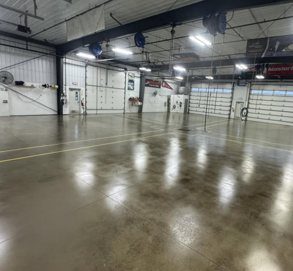Empty indoor garage with high ceiling, polished concrete floor, large overhead garage doors, and various tools and equipment along the walls.