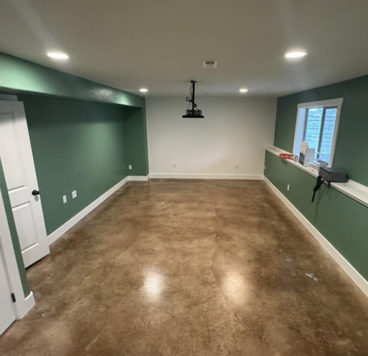 Empty room with green walls, hardwood floor, window, white door, and ceiling light fixtures, with some tools and supplies on the window sill.