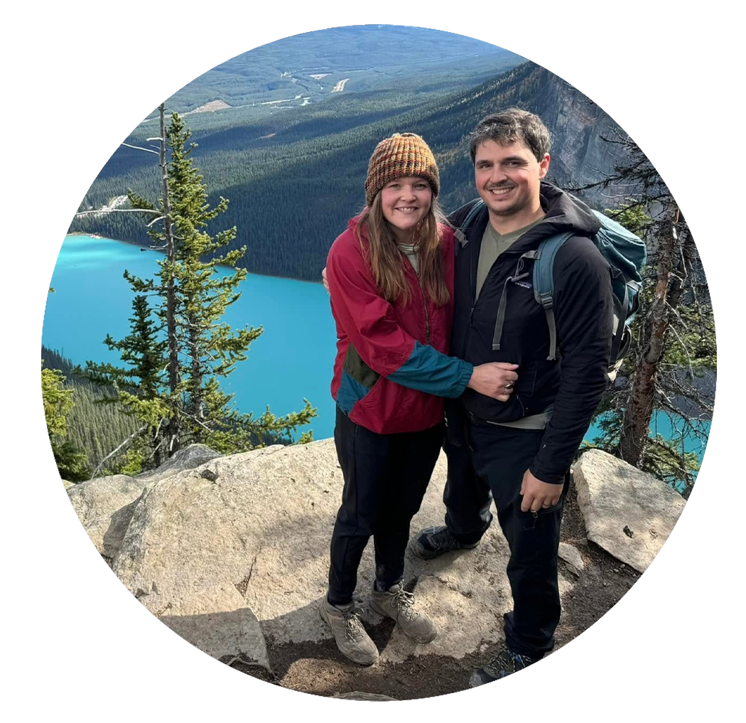 A couple hiking and smiling in front of a mountain lake, with trees and mountain landscape in the background.