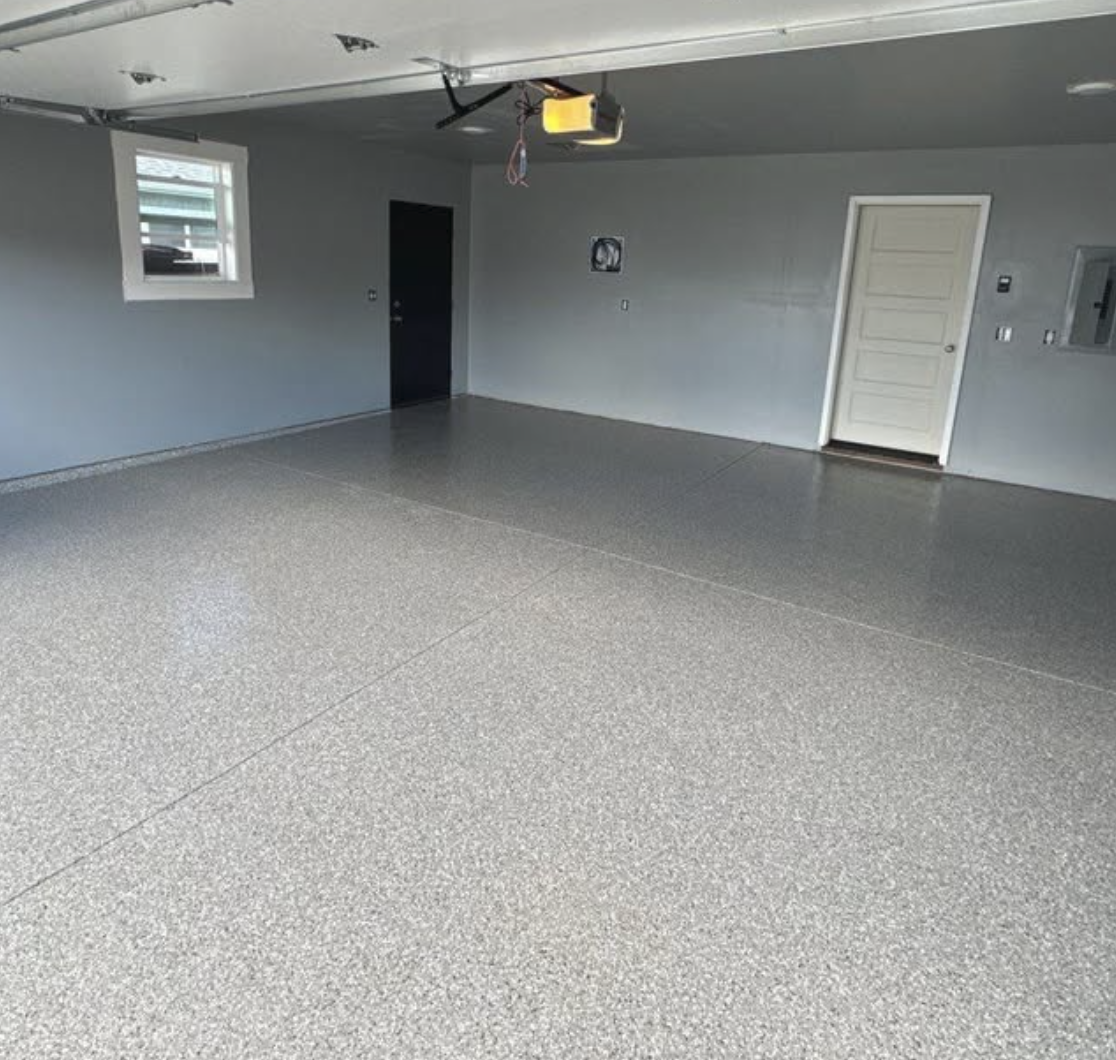 Empty garage with speckled gray epoxy floor, light gray walls, small window, door, and garage opener on ceiling.