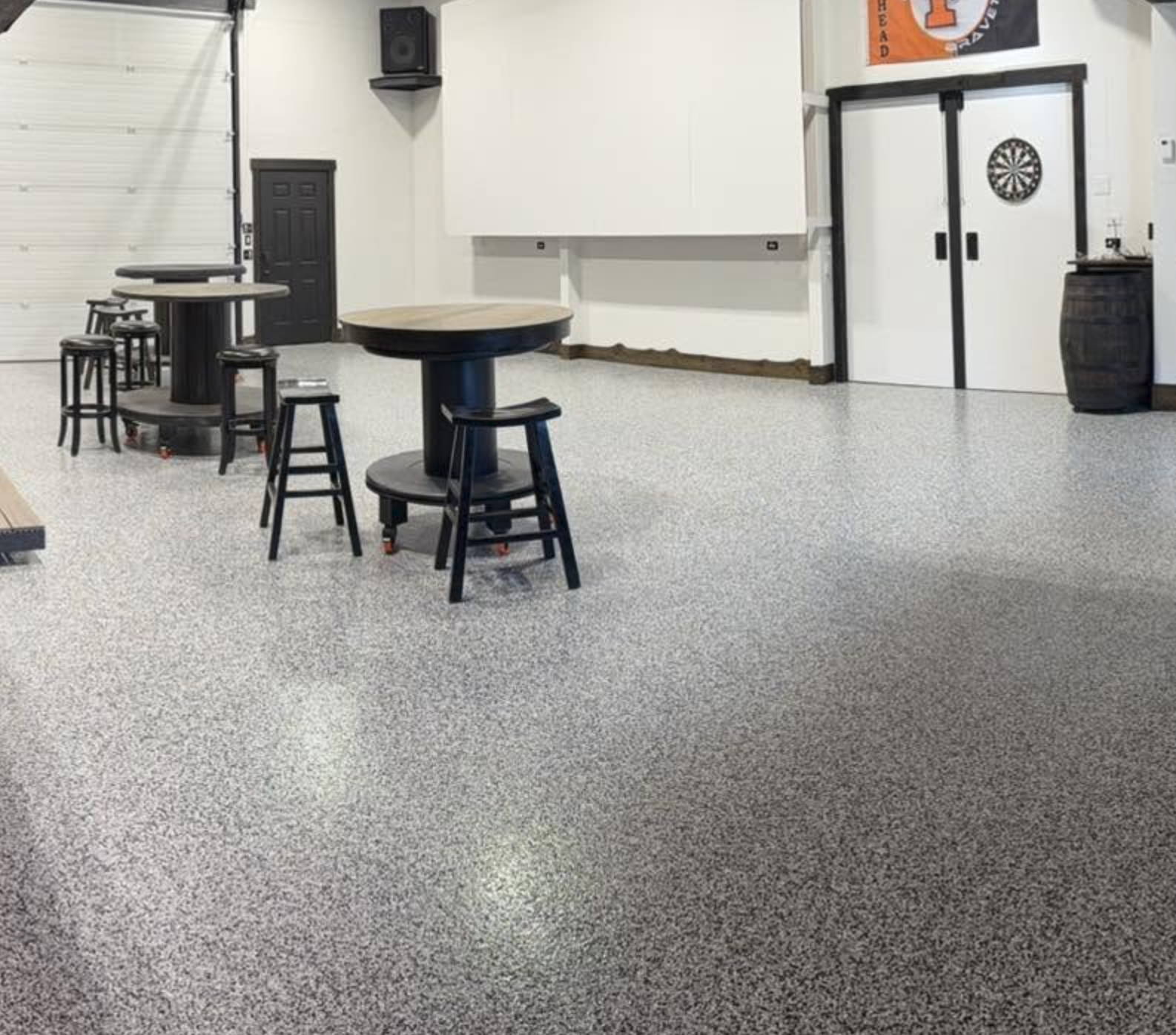 An indoor recreational space with two tables, several barstools, a dartboard on a white wall, a black door, a large garage door, and a Tennessee Volunteers banner.