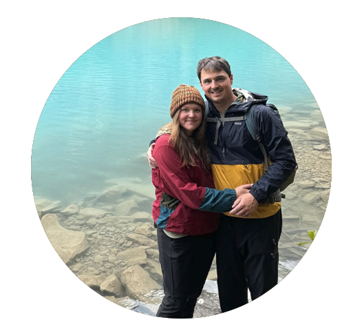 A couple standing together by a lake, smiling, with rocks and water in the background.
