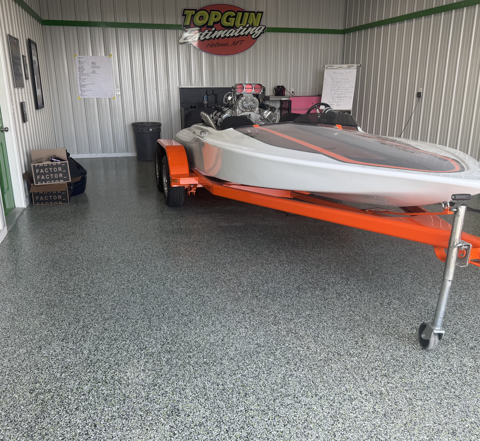 An indoor space with metal paneled walls and green trim, featuring a speedboat on a trailer, a poster with handwritten notes, and a sign reading 'Top Gun Estimating Helena, MT' hanging on the wall.