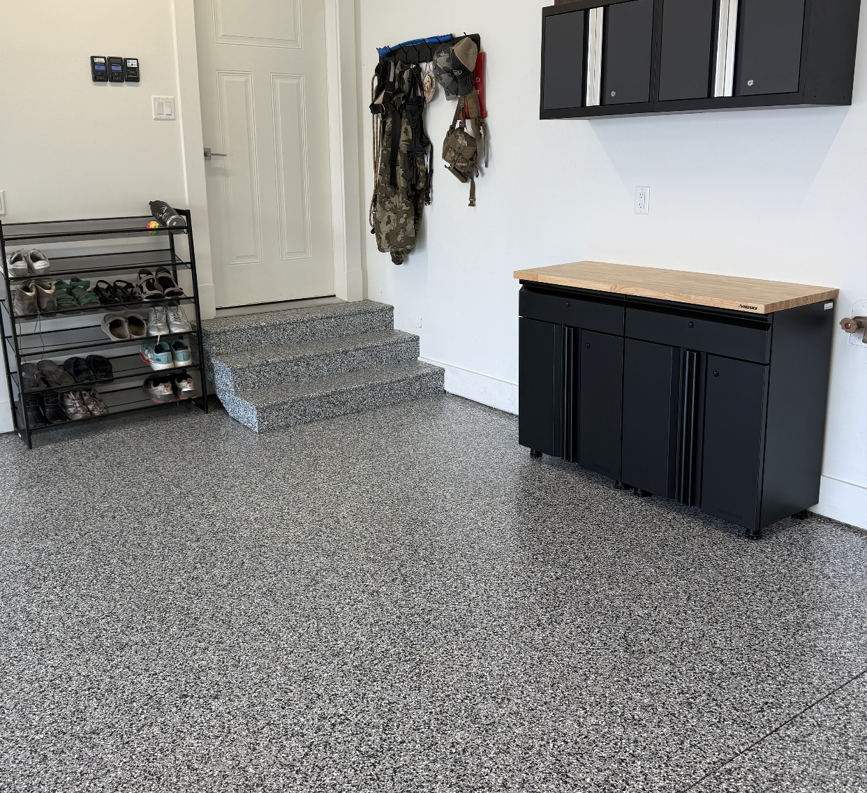 Entryway with shoe rack containing multiple pairs of shoes, a wall-mounted coat and hat rack with various gear, a black storage cabinet with a wooden top, and black wall-mounted cabinets. The flooring is speckled gray.