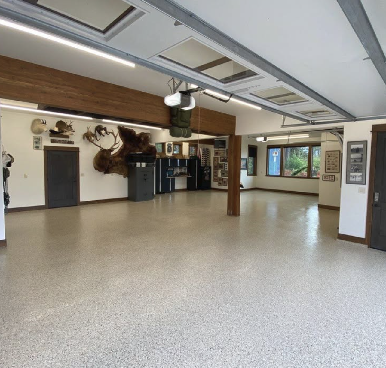 A spacious room with beige flooring, white walls, and large windows showing green trees outside. The room has wood accents, including a thick wooden beam and framed ceilings, with some mounted animal heads and framed photographs or artwork on the wal