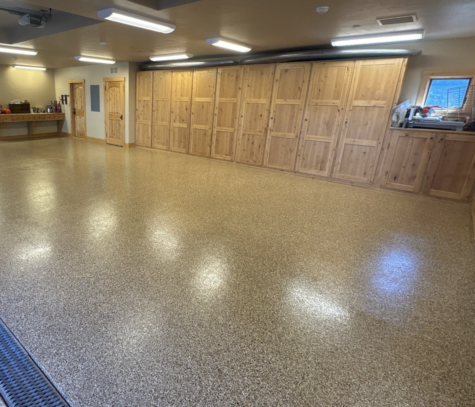 An empty room with beige speckled epoxy flooring, wooden cabinets along one wall, a small window, and ceiling lights.