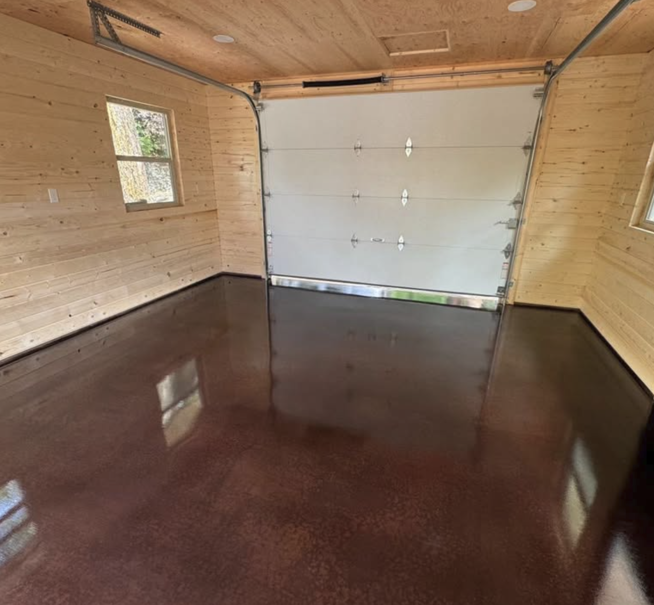 Empty garage with polished reddish-brown concrete floor, wooden walls, and a metal garage door with small windows. Two small windows on sides providing natural light.
