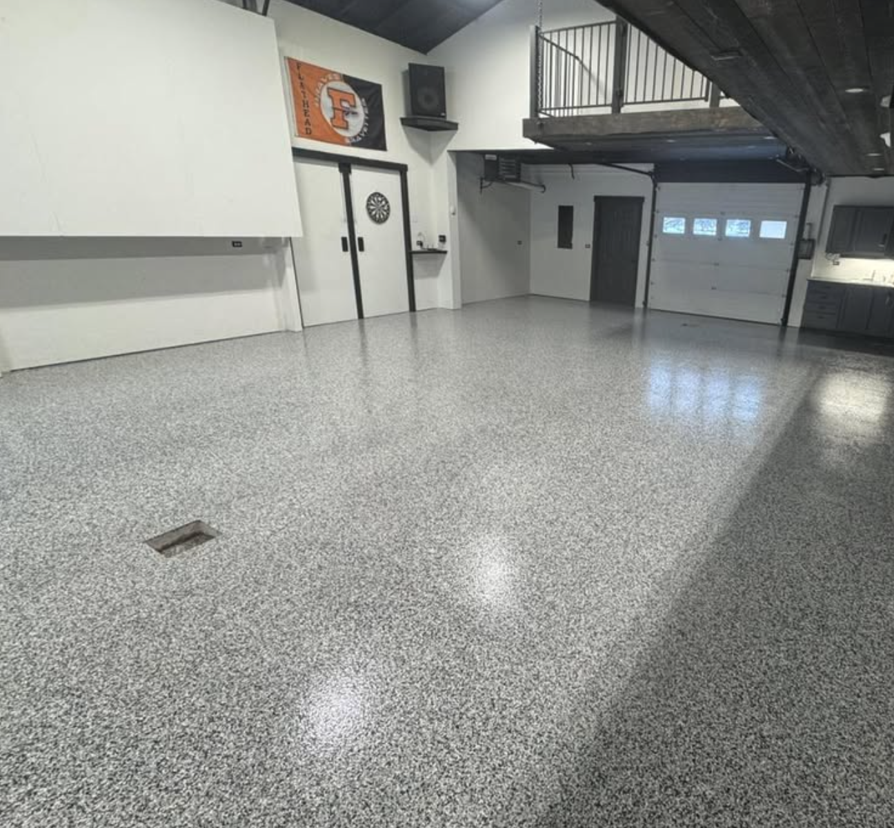 Empty garage with polished epoxy flooring, white walls, and a black ceiling, featuring a dartboard on the wall, a mounted speaker, and a staircase leading to a loft area.