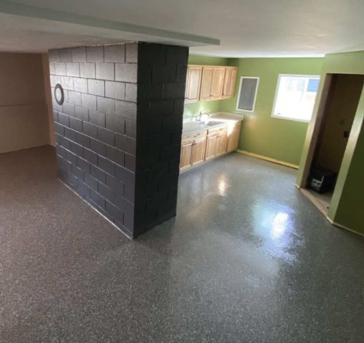 Interior view of a small kitchen with green walls, wooden cabinets, a window, and a black brick wall in the room.