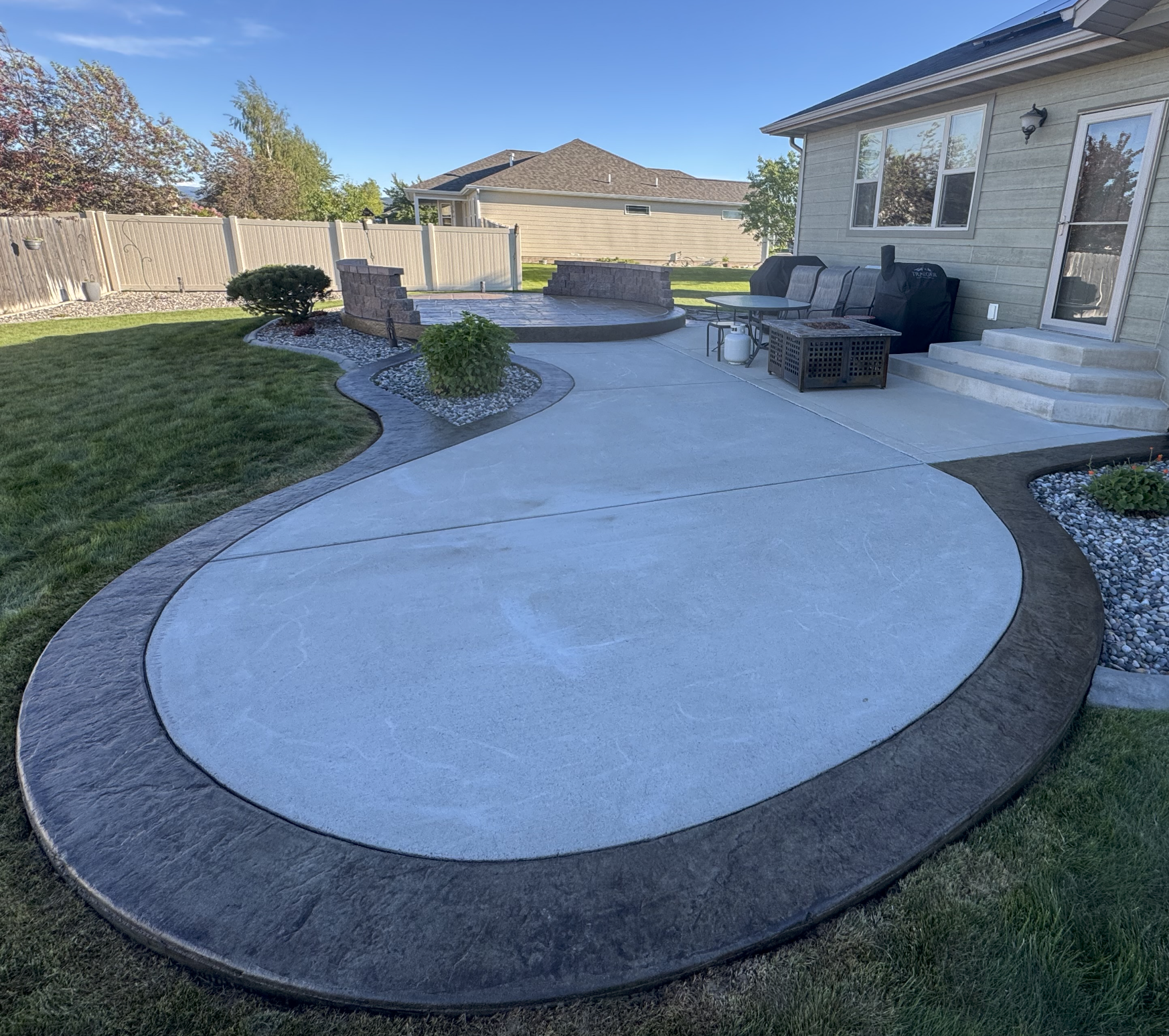 A backyard patio with a circular, textured concrete surface, surrounded by a curved border. There is a small landscaped area with a bush and decorative stones. The patio has steps leading to the house, which has windows and a door, with outdoor furni