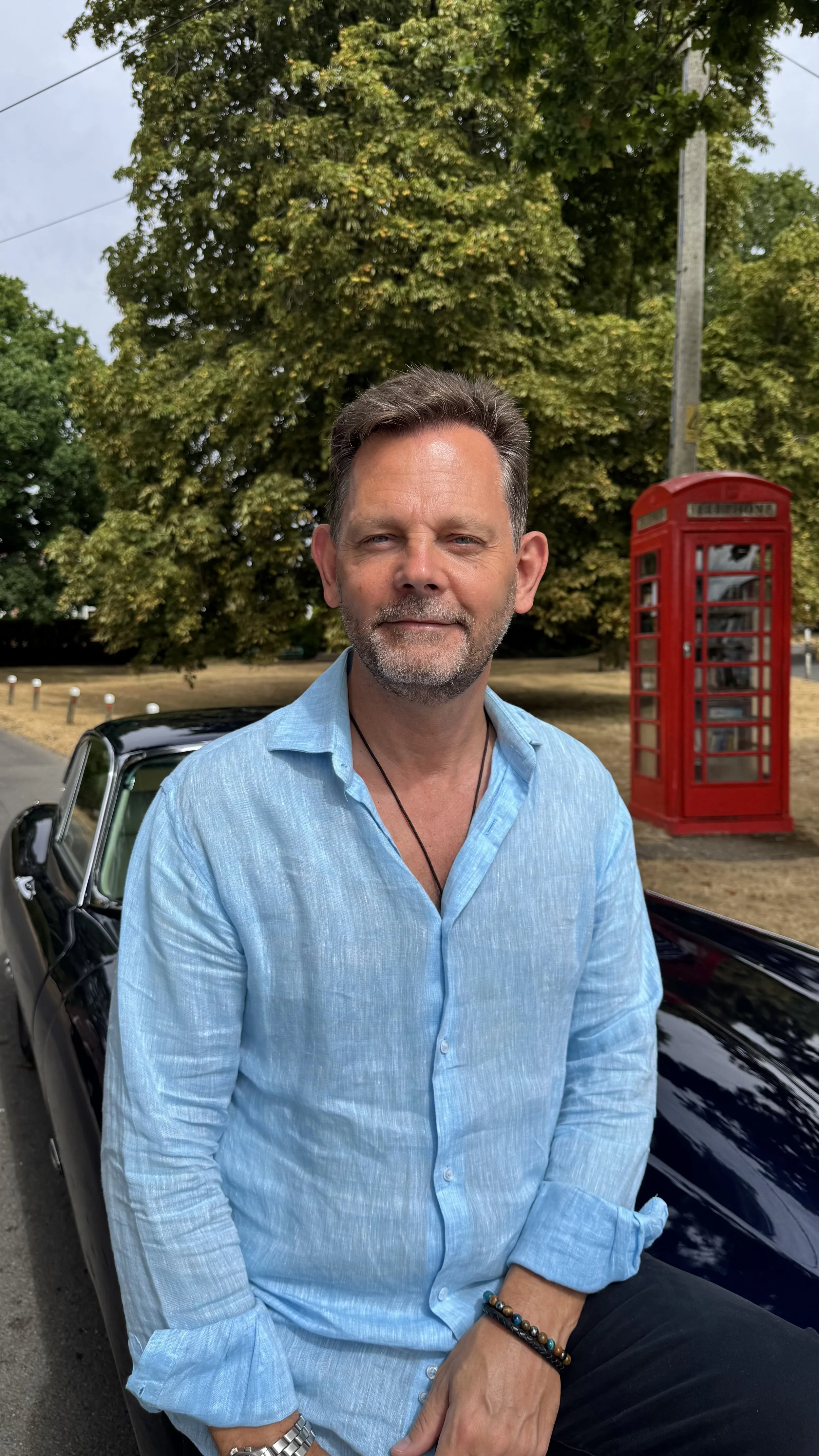 Paul Beach, founder of DriveWithPaul, sitting beside his 1971 Jaguar E-Type on the village green.