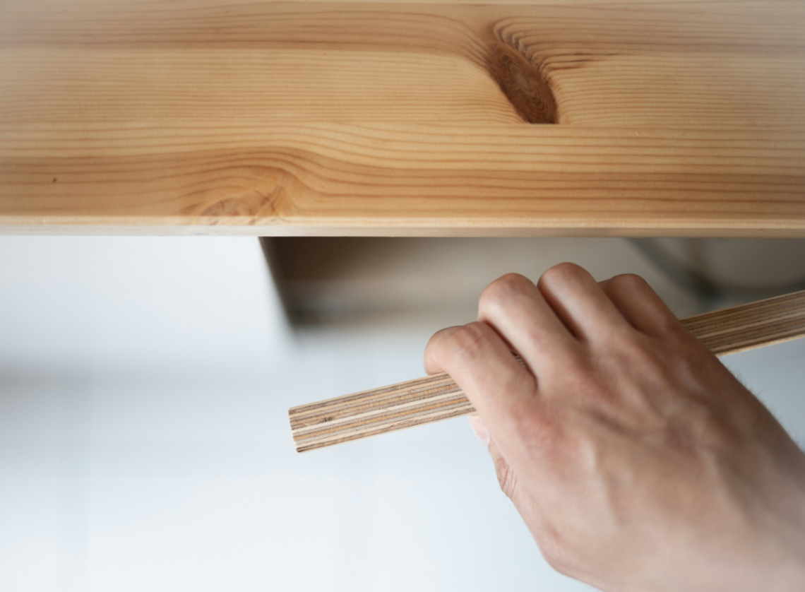 A person's hand holding a thin piece of wood under a larger wooden surface, likely a table or shelf, with visible wood grain.