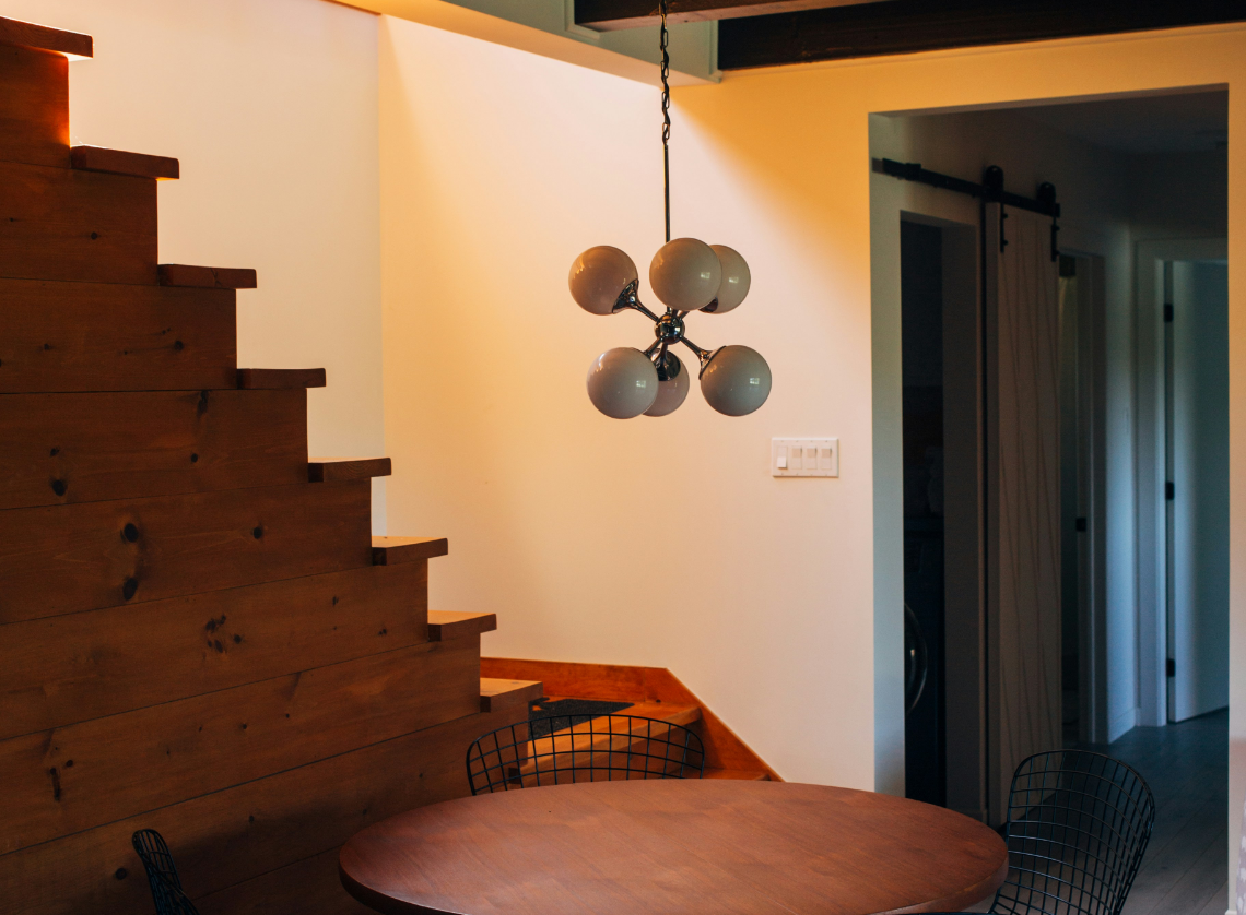 Interior view of a dining area with a round wooden table, black wire chairs, a wooden staircase, and a modern chandelier light fixture hanging from the ceiling.