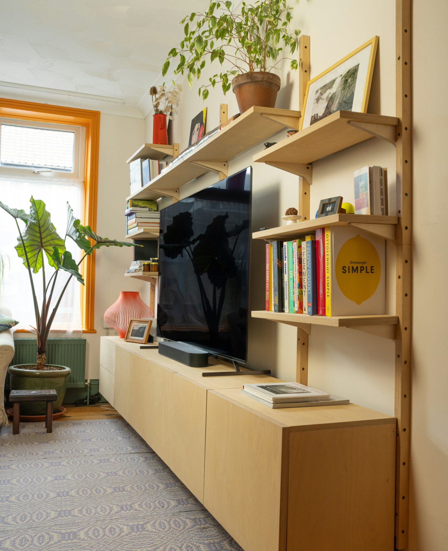 Living room with wooden wall-mounted shelves holding books, framed photos, and decorative items, a large flat-screen TV, a potted plant near a window with orange trim, a pink lamp, a piece of artwork, and a patterned carpet.