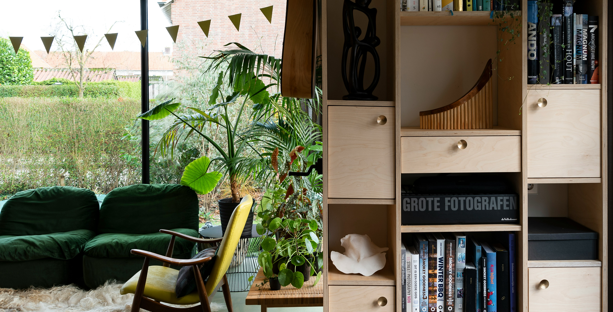 Custom plywood cabinetry and shelving in a warm, plant-filled interior with mid-century furniture.
