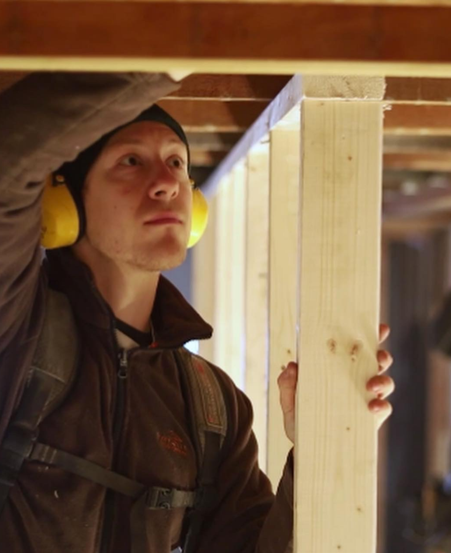 A man in a brown jacket and black hat is holding a piece of wood while working in a carpentry or woodworking shop.