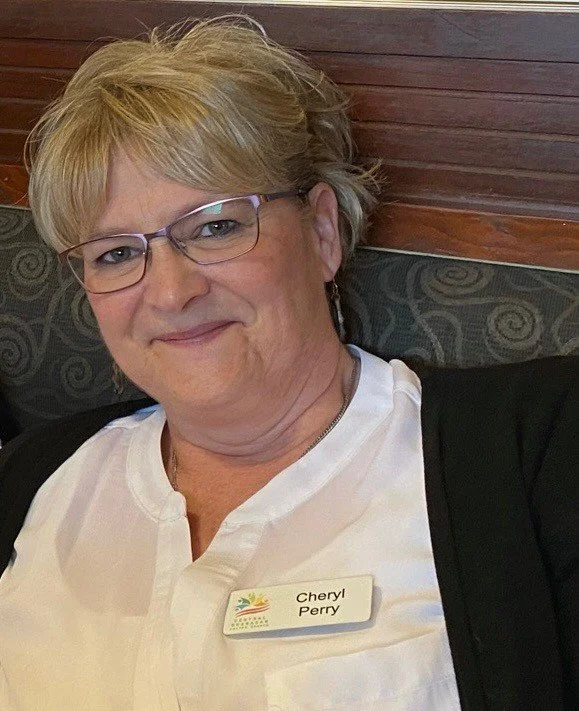 A woman with short blonde hair and glasses wearing a white blouse and a black cardigan, sitting at a restaurant, wearing a name tag that says Cheryl Perry.