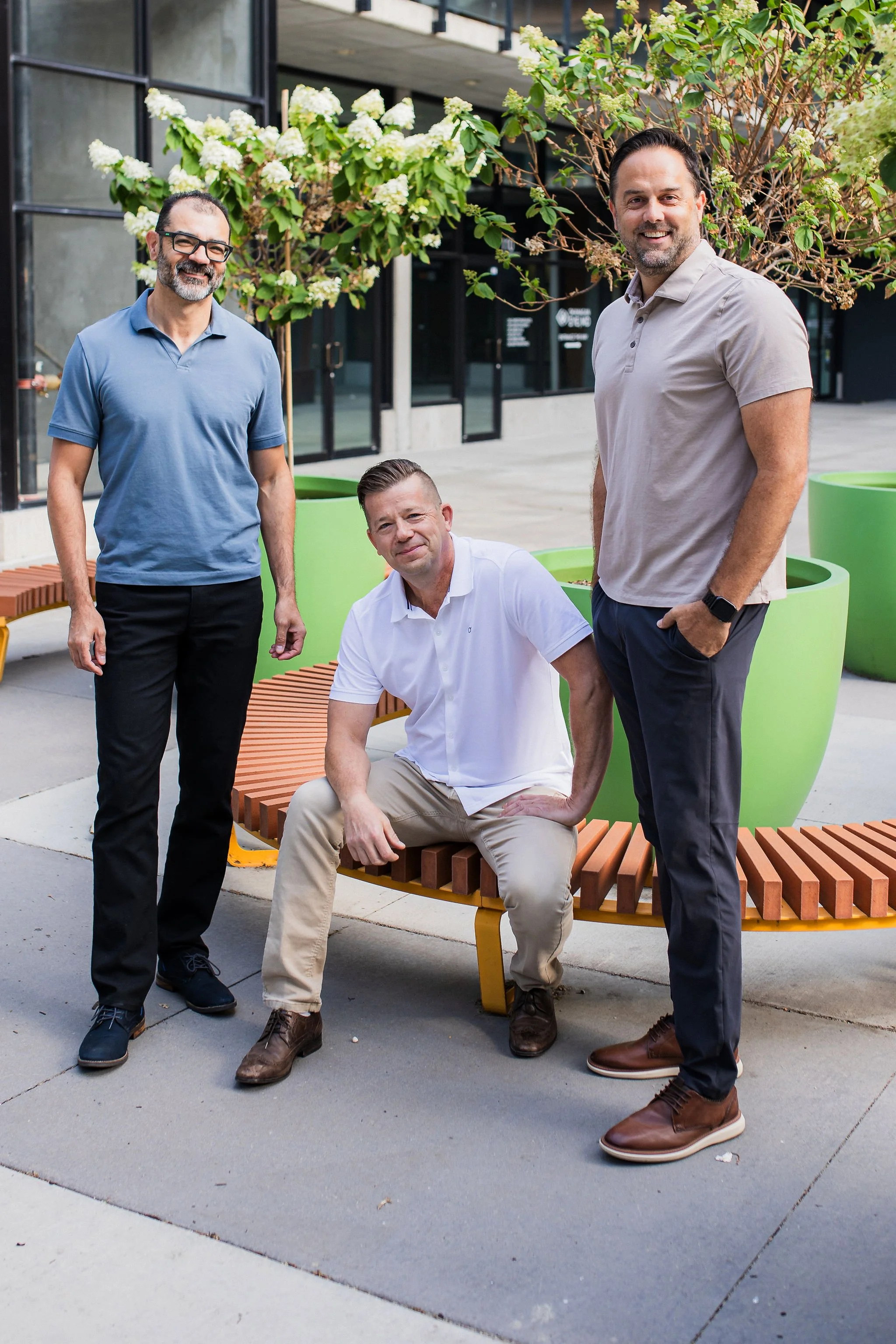 Three men in casual business attire outdoors on a sidewalk with benches and large green planters, posing for the photo.