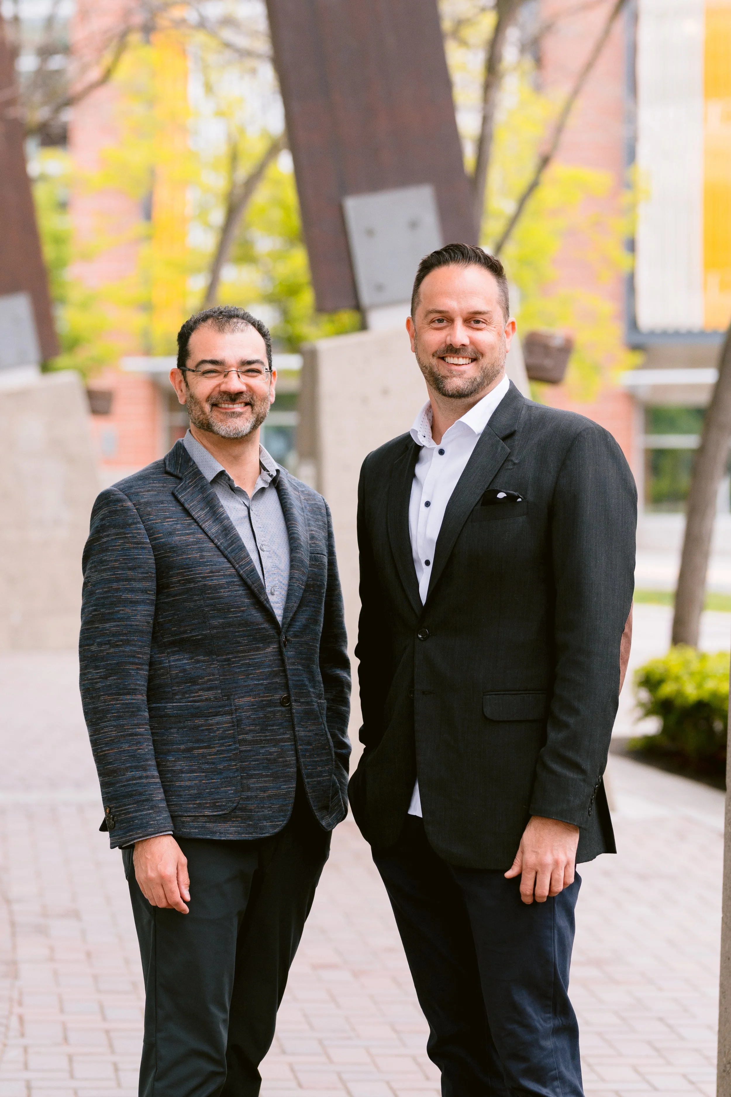 Two men in suits standing outdoors smiling for the camera with trees and building in the background.