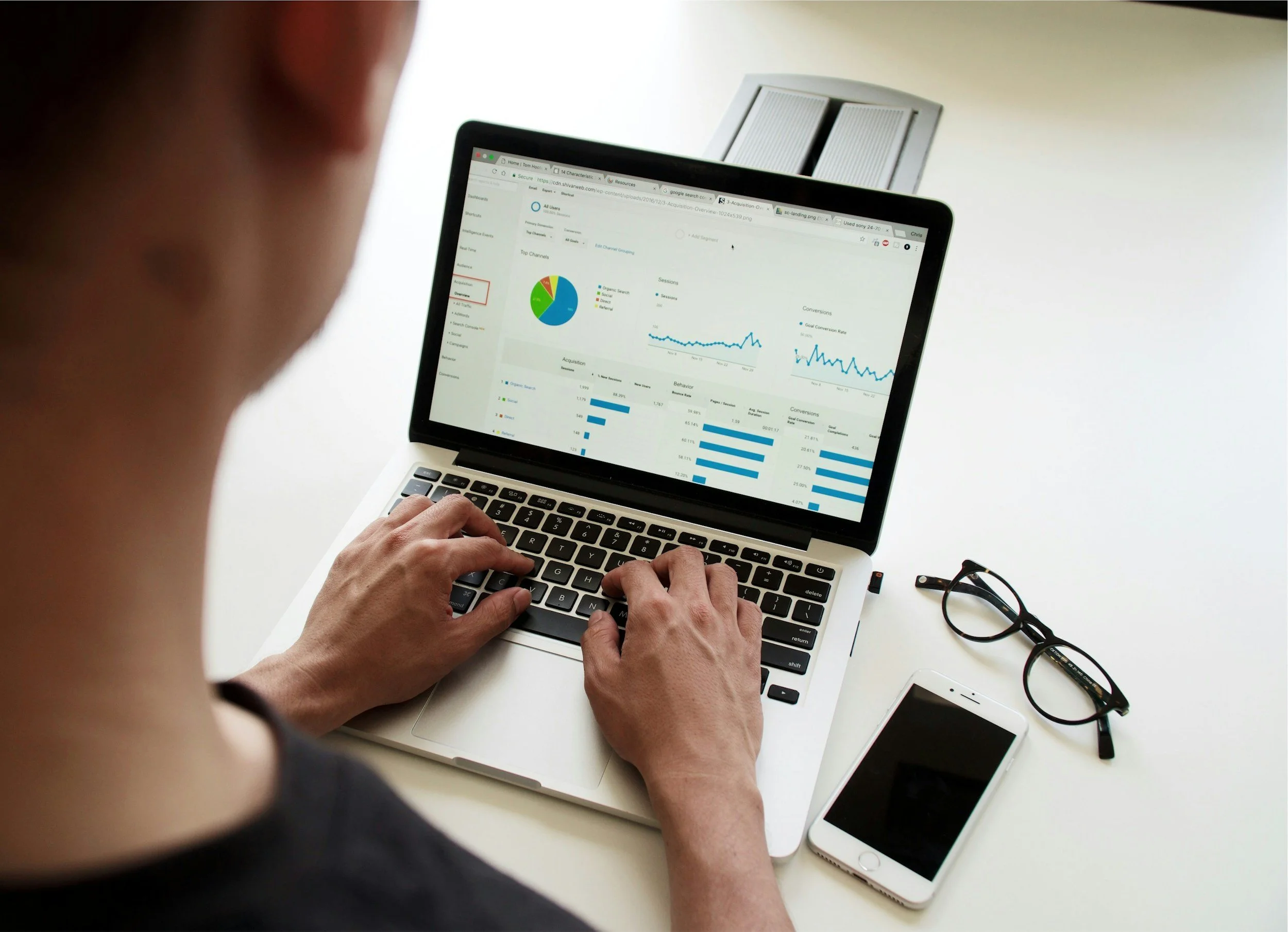 Person working on a laptop displaying analytics and graphs, with a smartphone, glasses, and a pen on a white table.