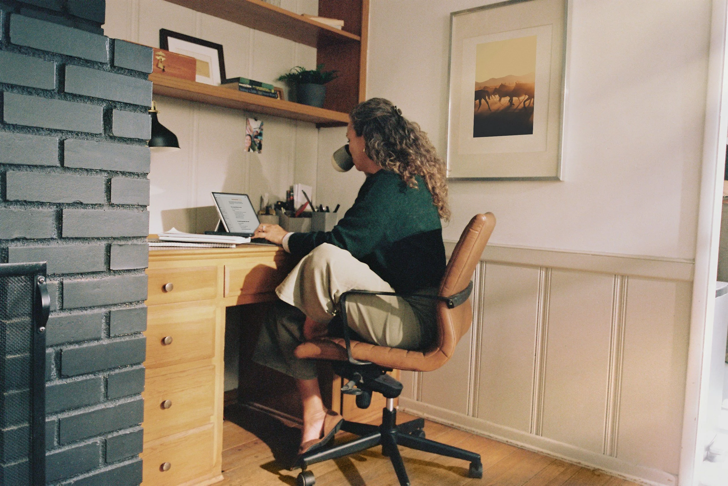 A woman with curly hair sitting at a wooden desk using a tablet computer, in a cozy room with shelves, framed artwork, and a large window.