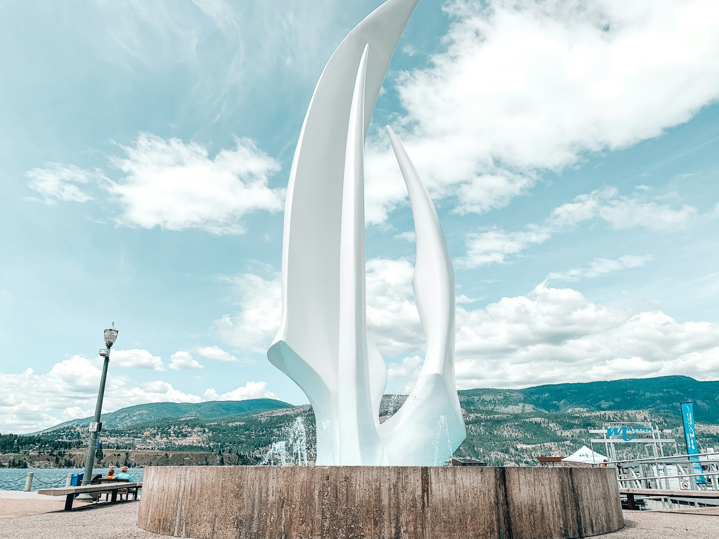 A large white modern sculpture resembling a wind turbine or abstract sail structures, set against a cloudy sky with mountains in the background, near a body of water with benches and streetlamps in the foreground.