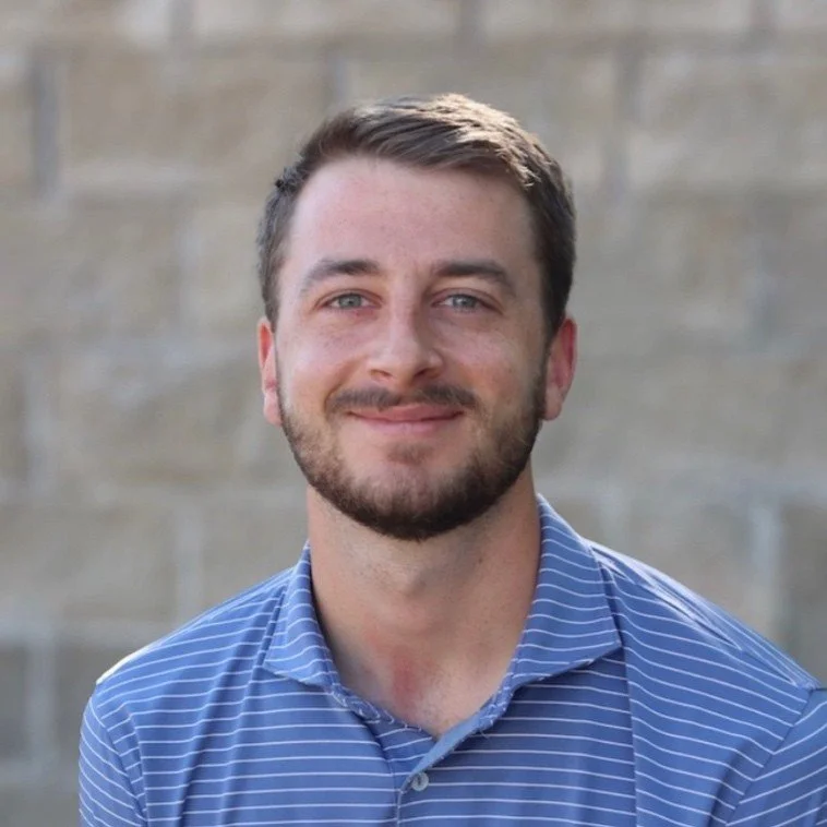 A young man with short brown hair and a beard, smiling, wearing a blue pinstripe collared shirt, against a grey brick wall background.