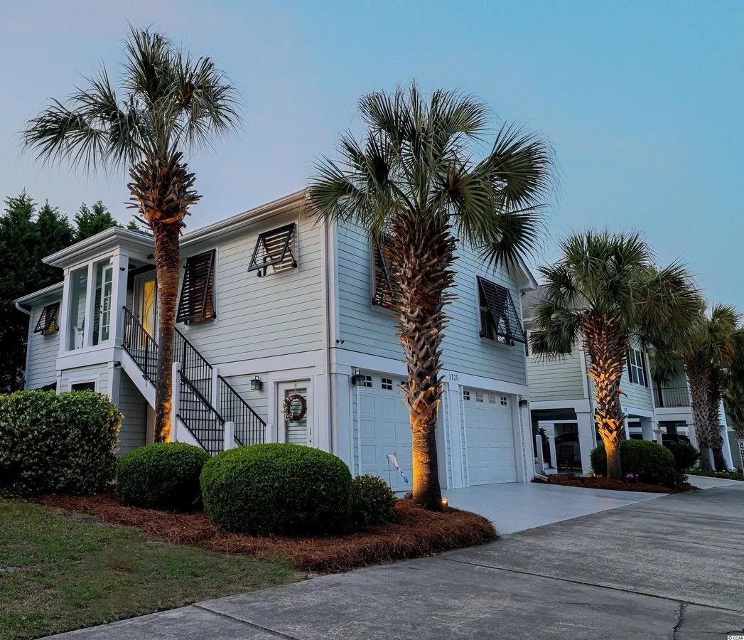 A row of modern white townhouses with garage doors, surrounded by well-manicured bushes and palm trees, at dusk.
