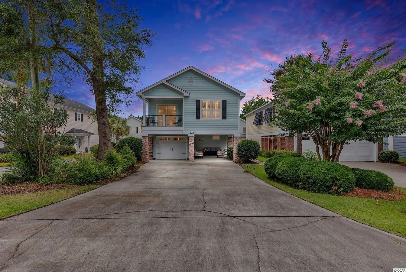 A two-story house with a blue exterior, black shutters, brick pillars, and a small front porch, surrounded by greenery and trees under a colorful sunset sky.