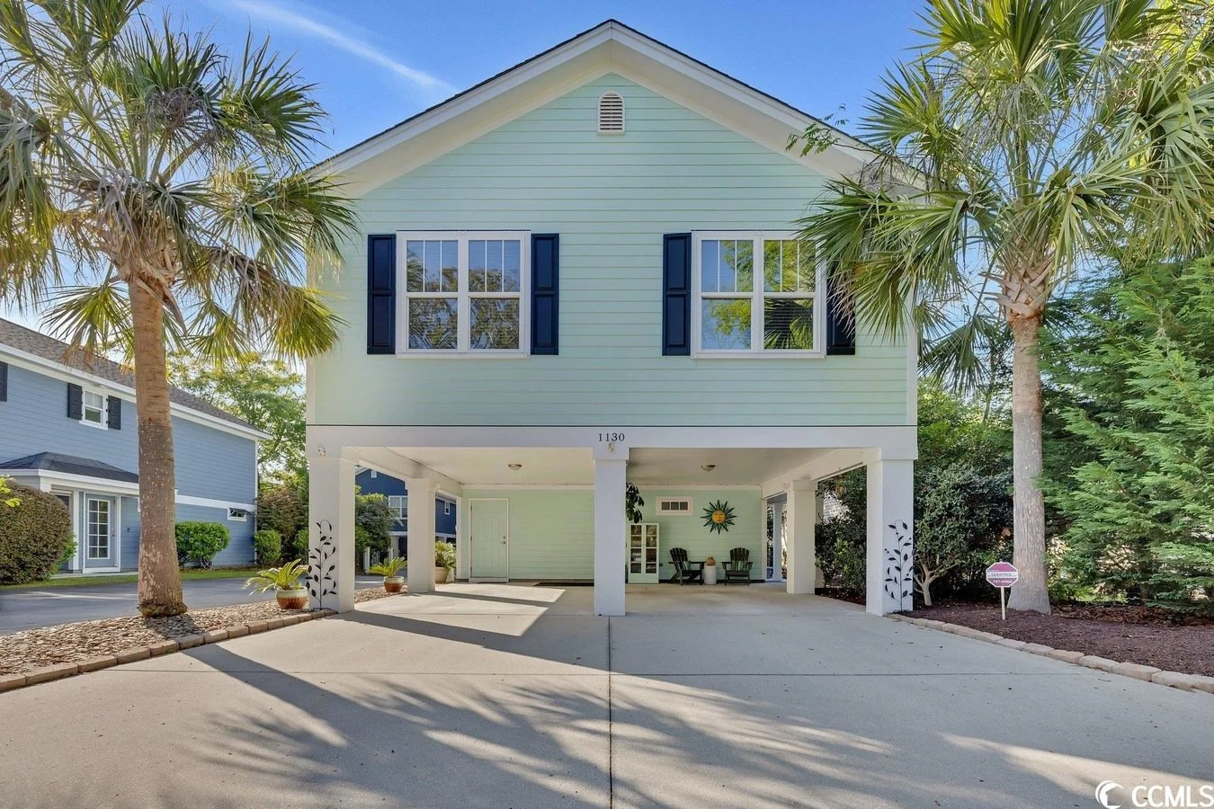 A two-story house painted in light pastel green with dark blue window shutters, situated in a tropical setting with palm trees, a tidy driveway, and a covered parking area decorated with potted plants and outdoor chairs.