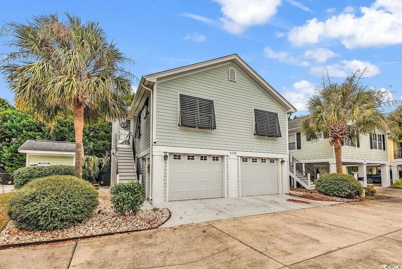 A two-story house with three garage doors, surrounded by palm trees and bushes, under a partly cloudy sky.