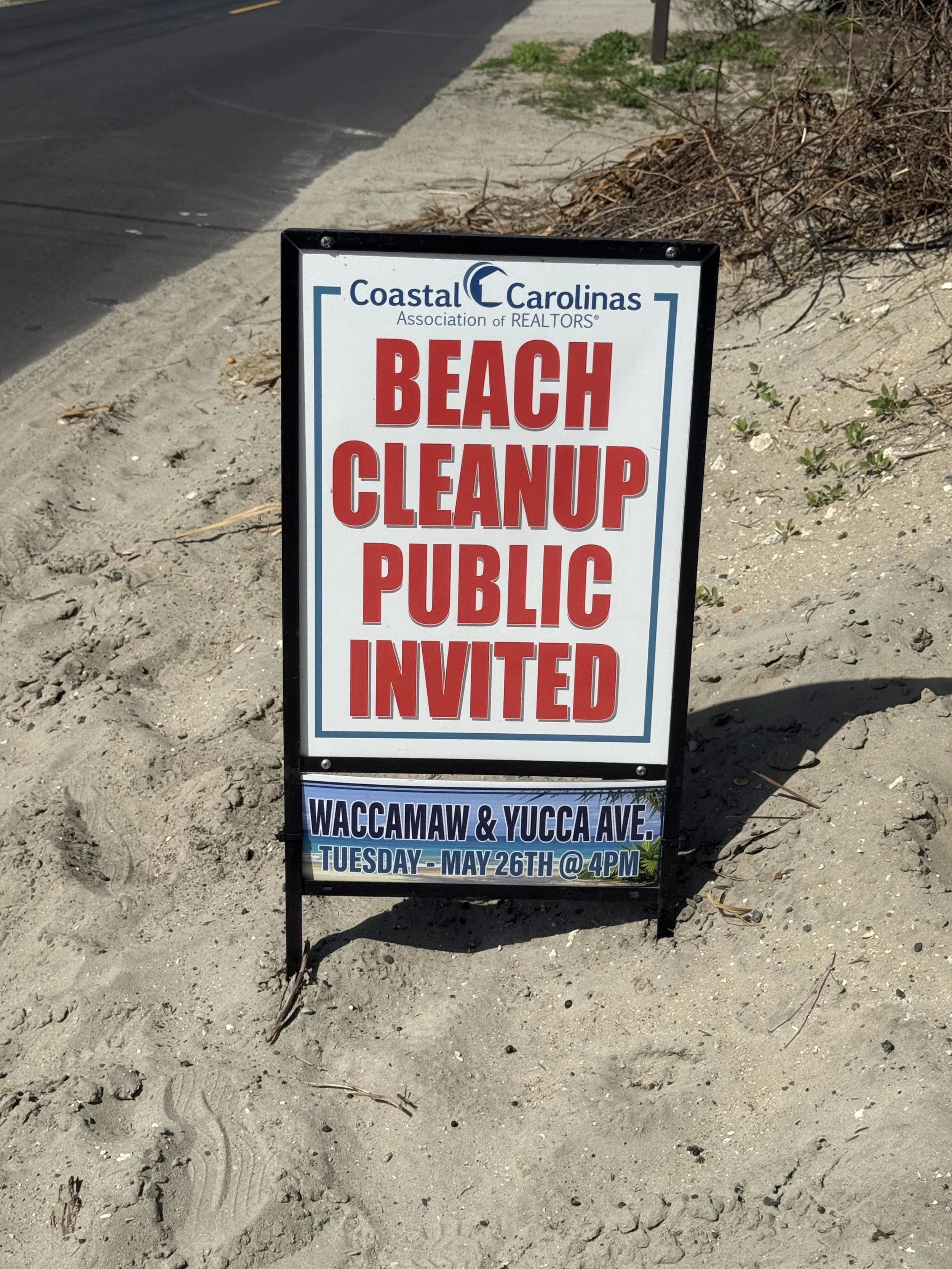 Signboard on the sandy beach area reads 'Coastal Carolinas Association of REALTORS, Beach Cleanup Public Invited, Waccamaw & YUCCA Ave, Tuesday-May 26th @ 4PM' with some green plants and dried twigs around.