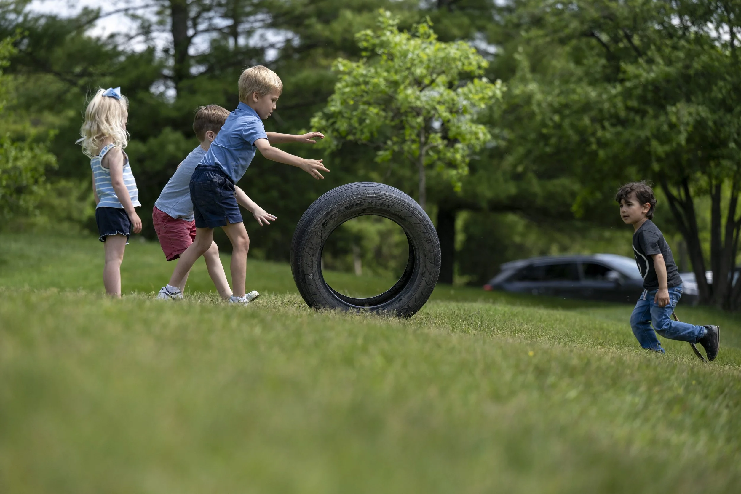 Four children playing outdoors on a grassy field with trees in the background, passing a large tire.