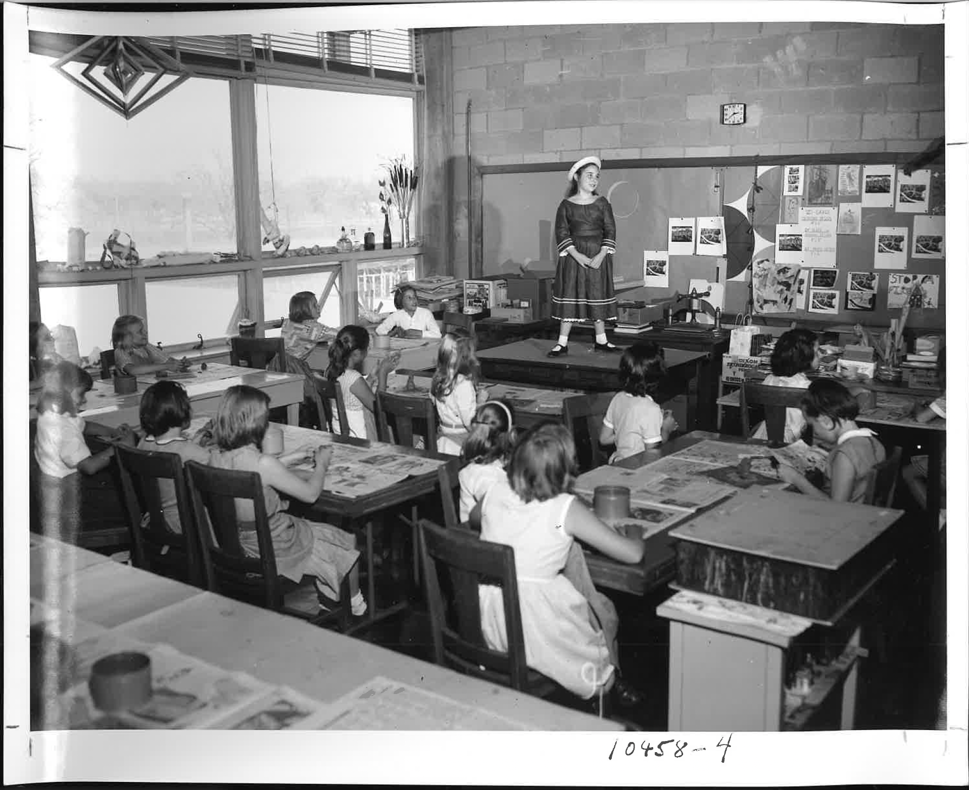 A classroom with students sitting at desks, listening to a female teacher standing on a small stage. The classroom has large windows with plants and decorations. The wall behind the teacher features educational posters and a clock.