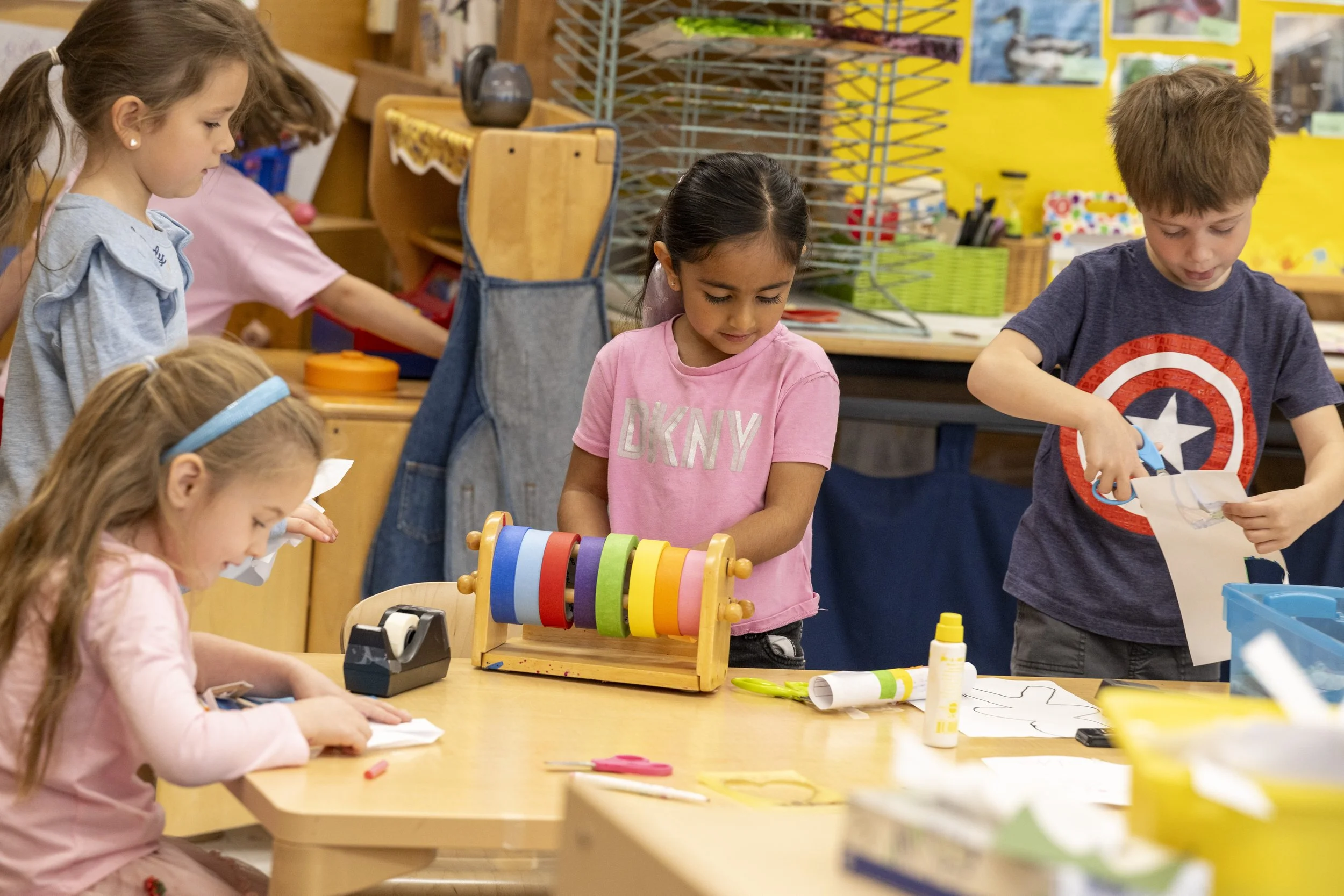 Children working on arts and crafts at a classroom table with art supplies and a colorful toy.