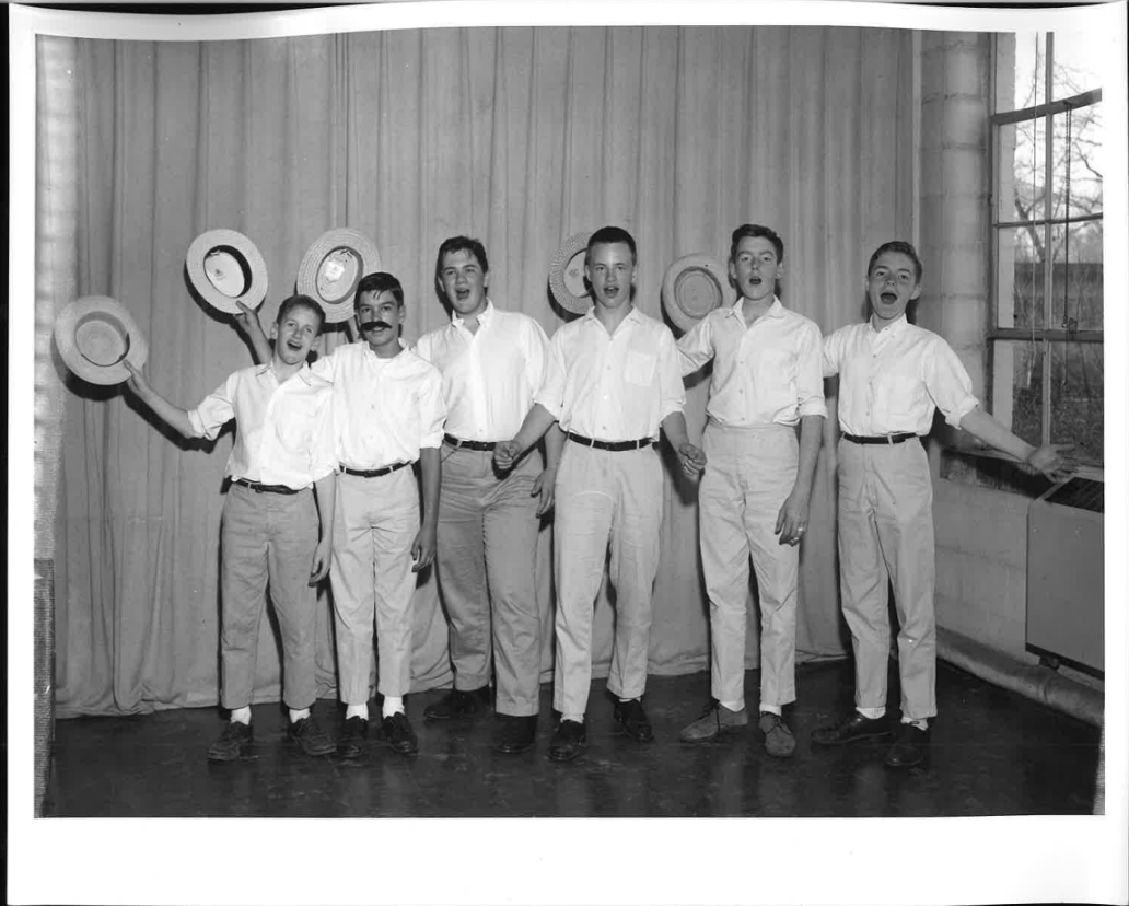 Seven young men wearing white shirts and light pants, standing in a line inside a room with a wooden wall and a window, posing with hats in the air and smiling.