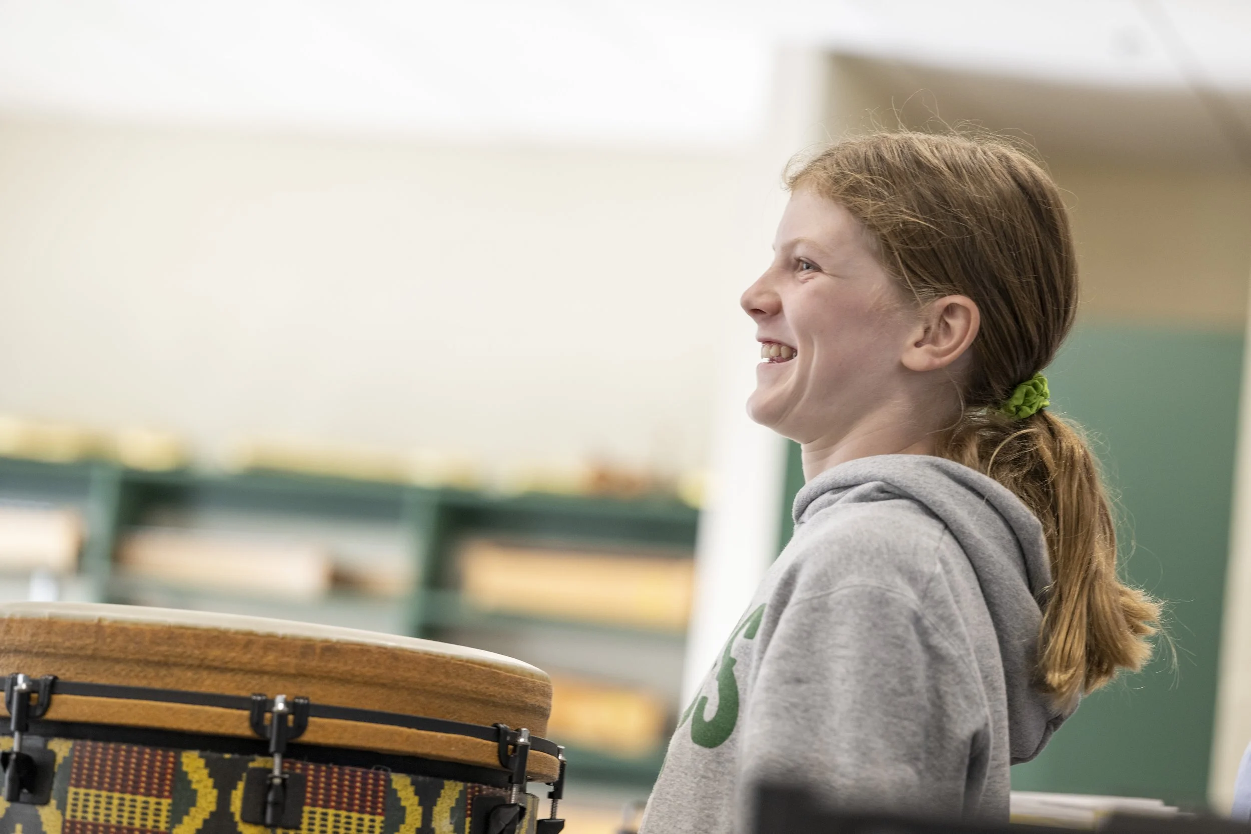 A girl with red hair tied with a green band wearing a gray hoodie smiling while playing a drum in a classroom or music room.