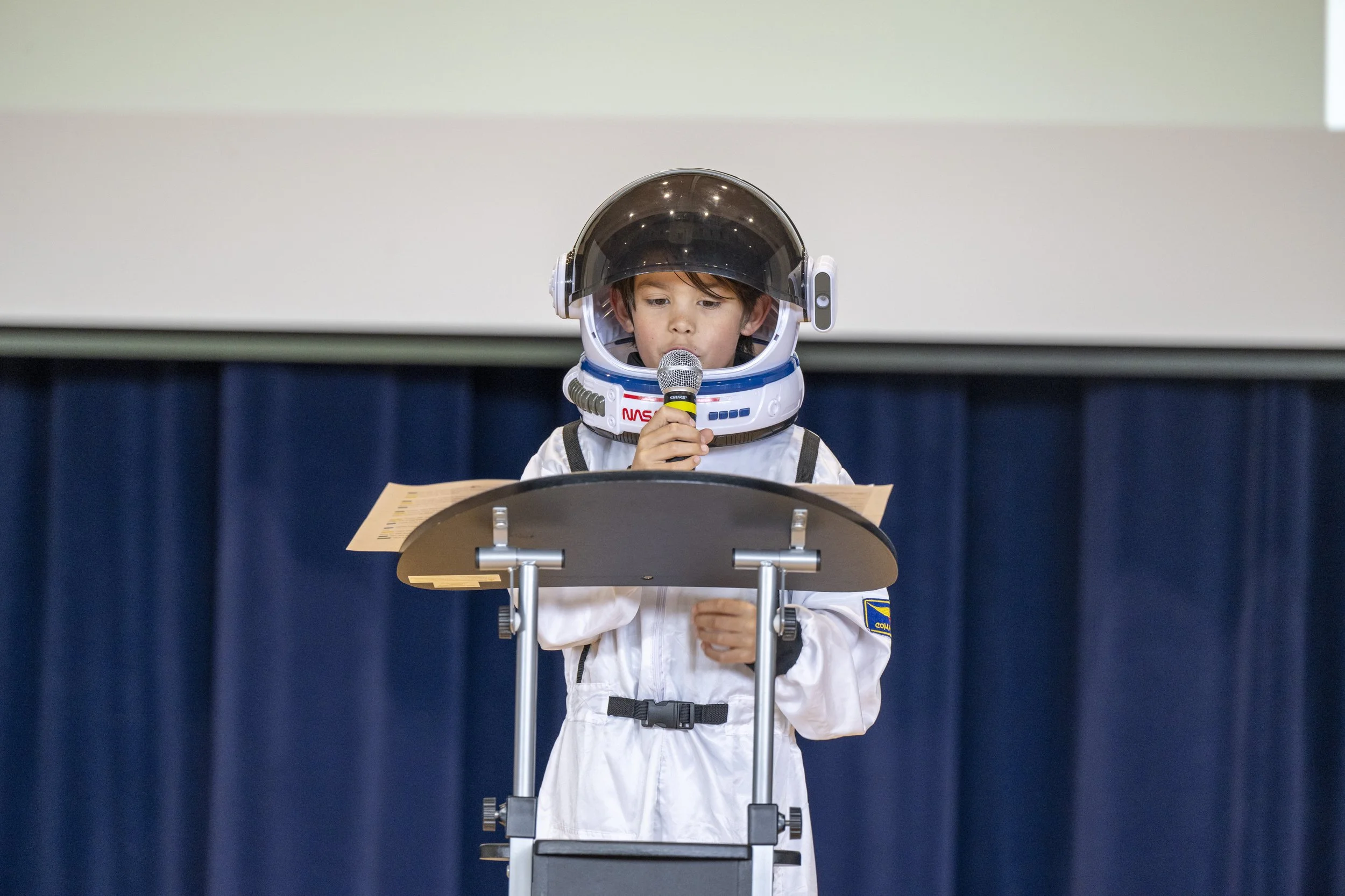Young boy dressed as an astronaut reading from a podium on a stage.