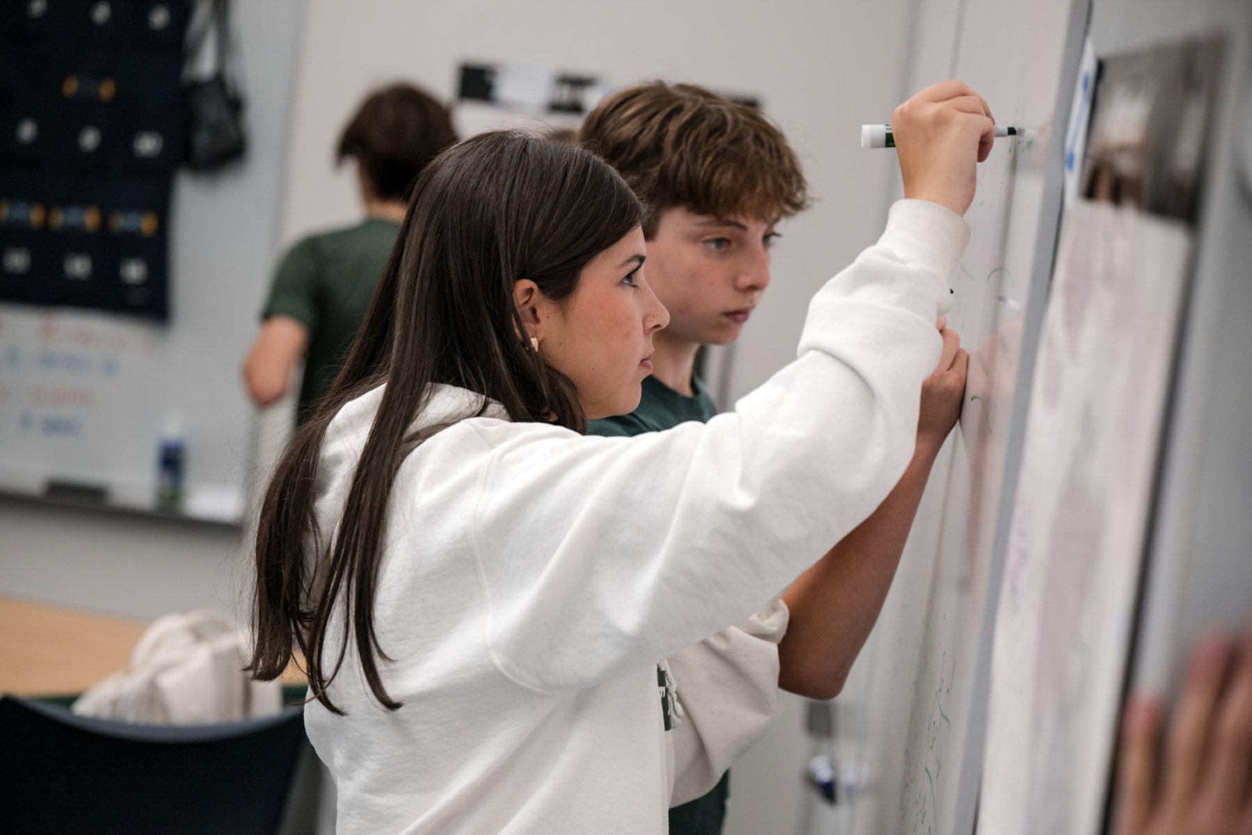 A girl writing on a whiteboard with a marker, with a boy standing next to her, in a classroom setting.