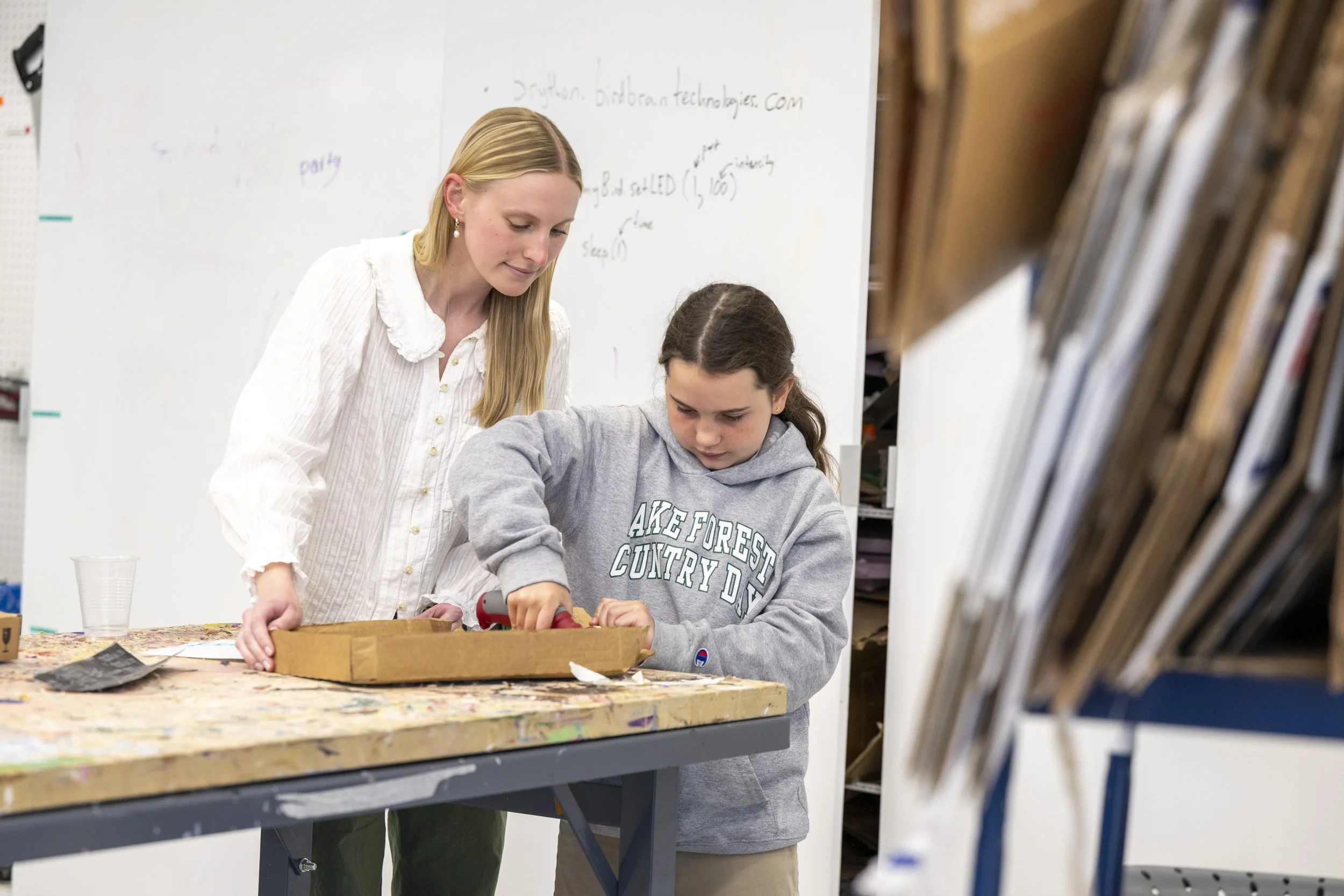 A woman and a girl are working together on a project at a table in an art classroom, with shelves of art supplies nearby and a whiteboard in the background.