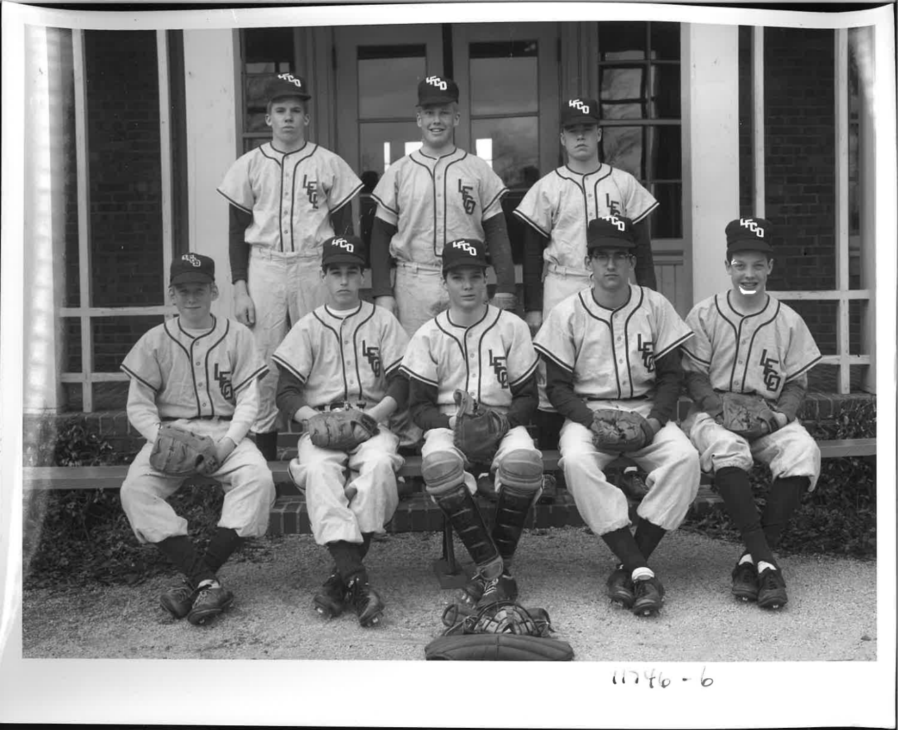 Black and white photograph of a vintage youth baseball team posing outdoors in front of a building with large windows. The team consists of nine boys in baseball uniforms and caps, some holding baseball gloves. They are arranged with four sitting on 