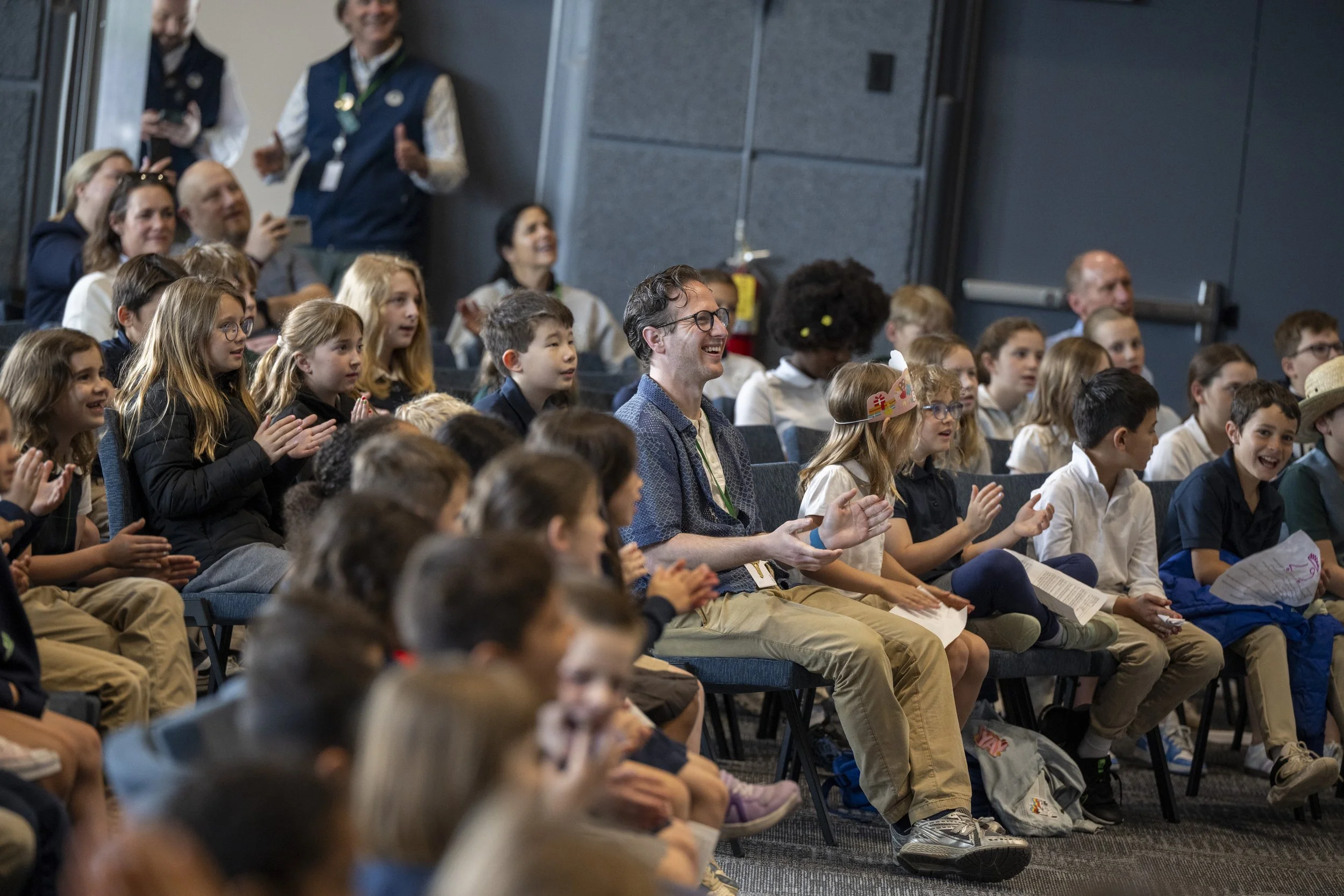 A group of children and adults seated in an auditorium, watching a presentation and applauding. Some are smiling and appear engaged.