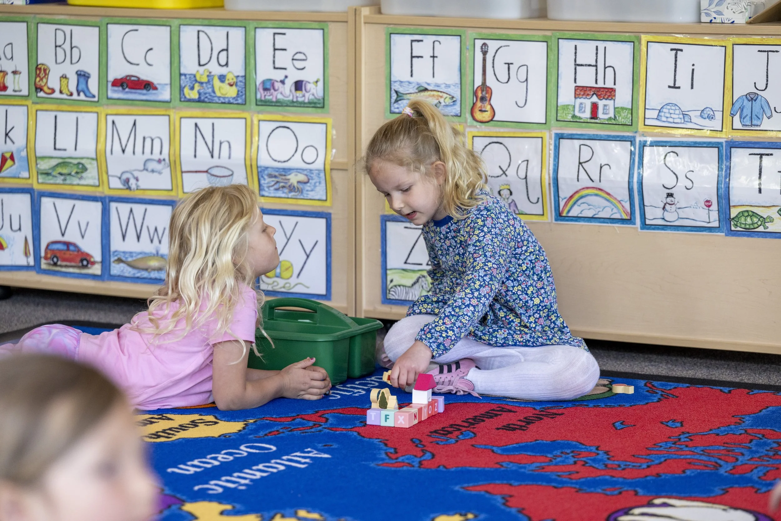 Two young girls are sitting on a colorful classroom rug, playing with blocks. The background features alphabet posters with illustrated images, including a house, fish, guitar, and boat.