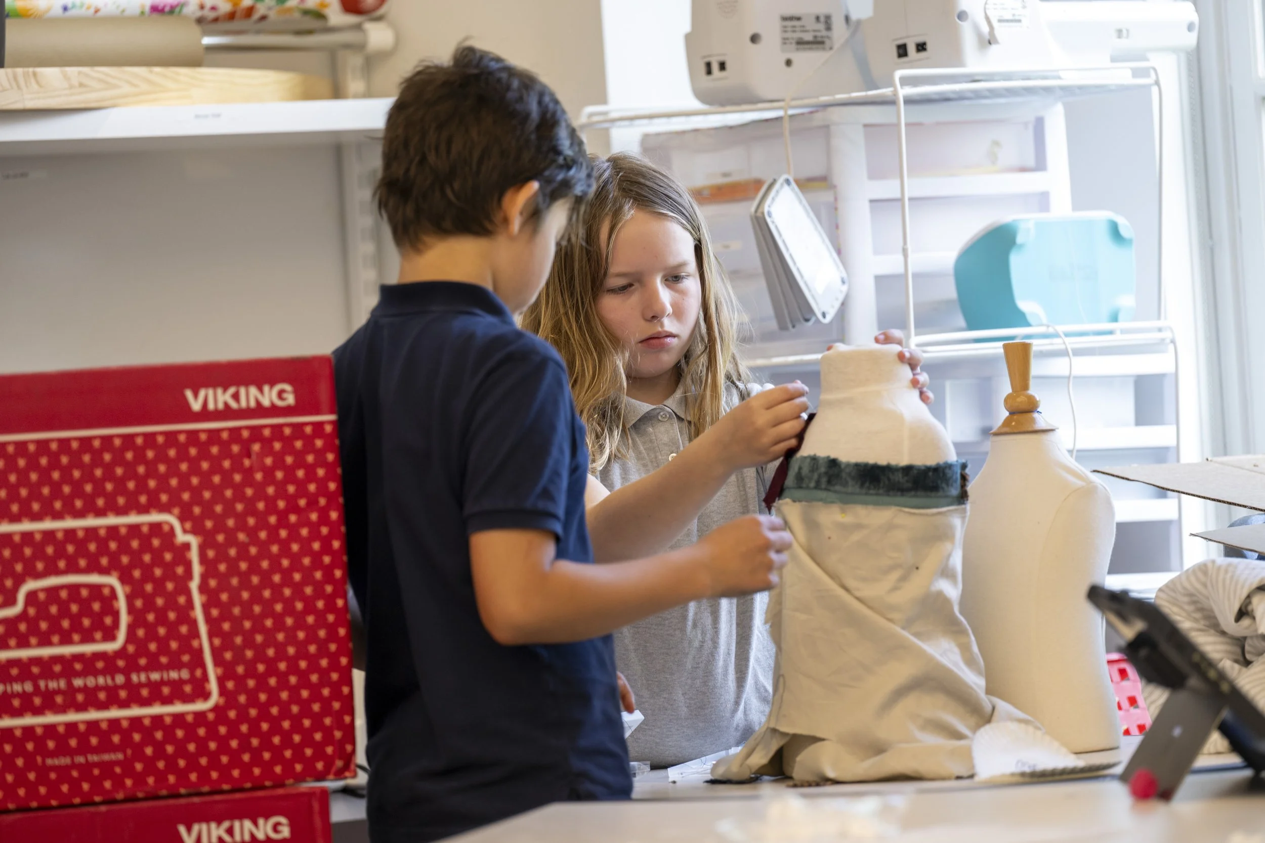 Two children working on a craft project involving fabric, with sewing supplies and a Viking sewing machine box visible in the background.