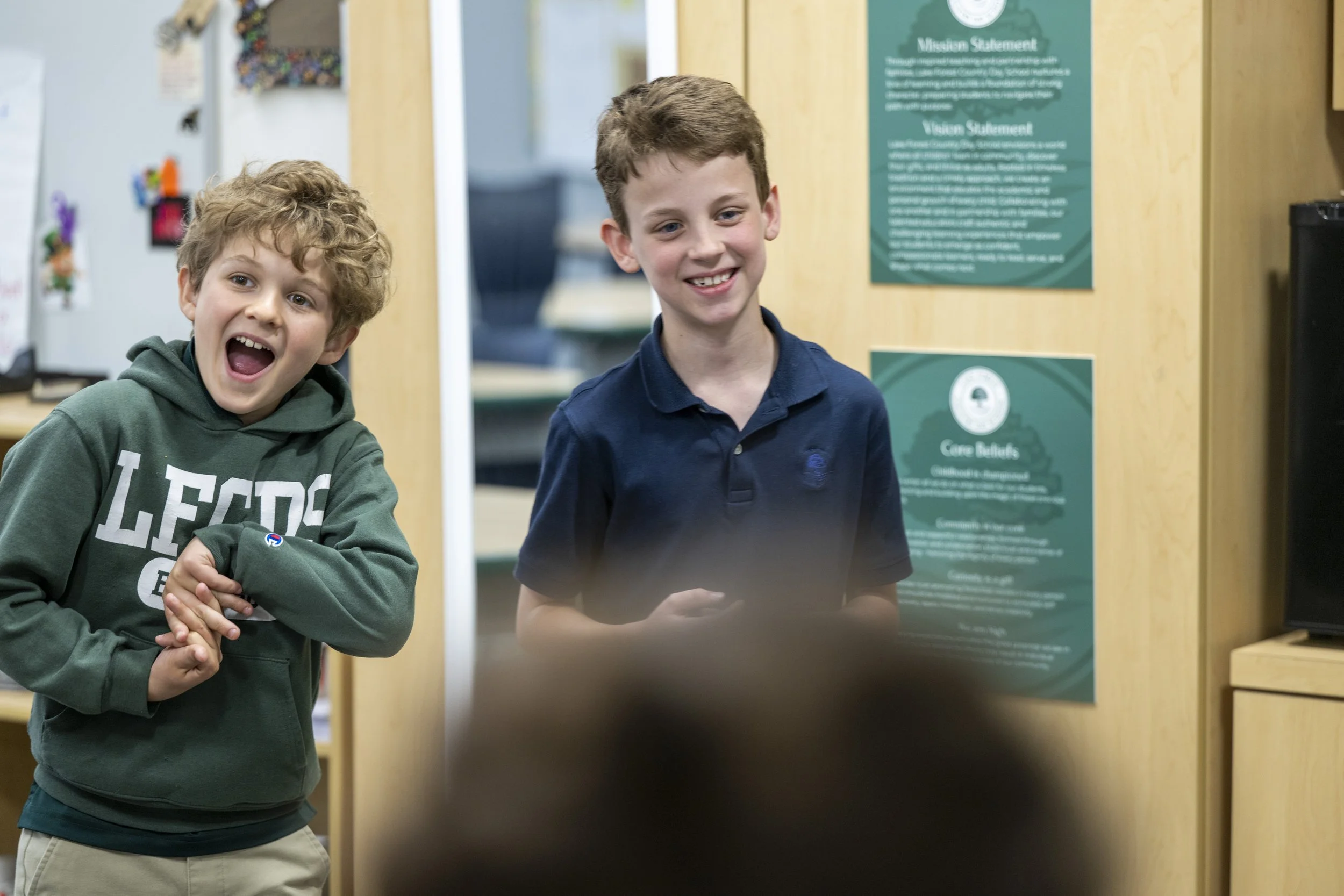 Two boys standing next to each other, smiling in a school or community center setting. The boy on the left has curly hair, wearing a green hoodie with white text, and the boy on the right has straight hair, wearing a navy polo shirt. Behind them is a wooden wall with green informational posters, including one titled 'Mission Statement' and another titled 'Core Beliefs'.