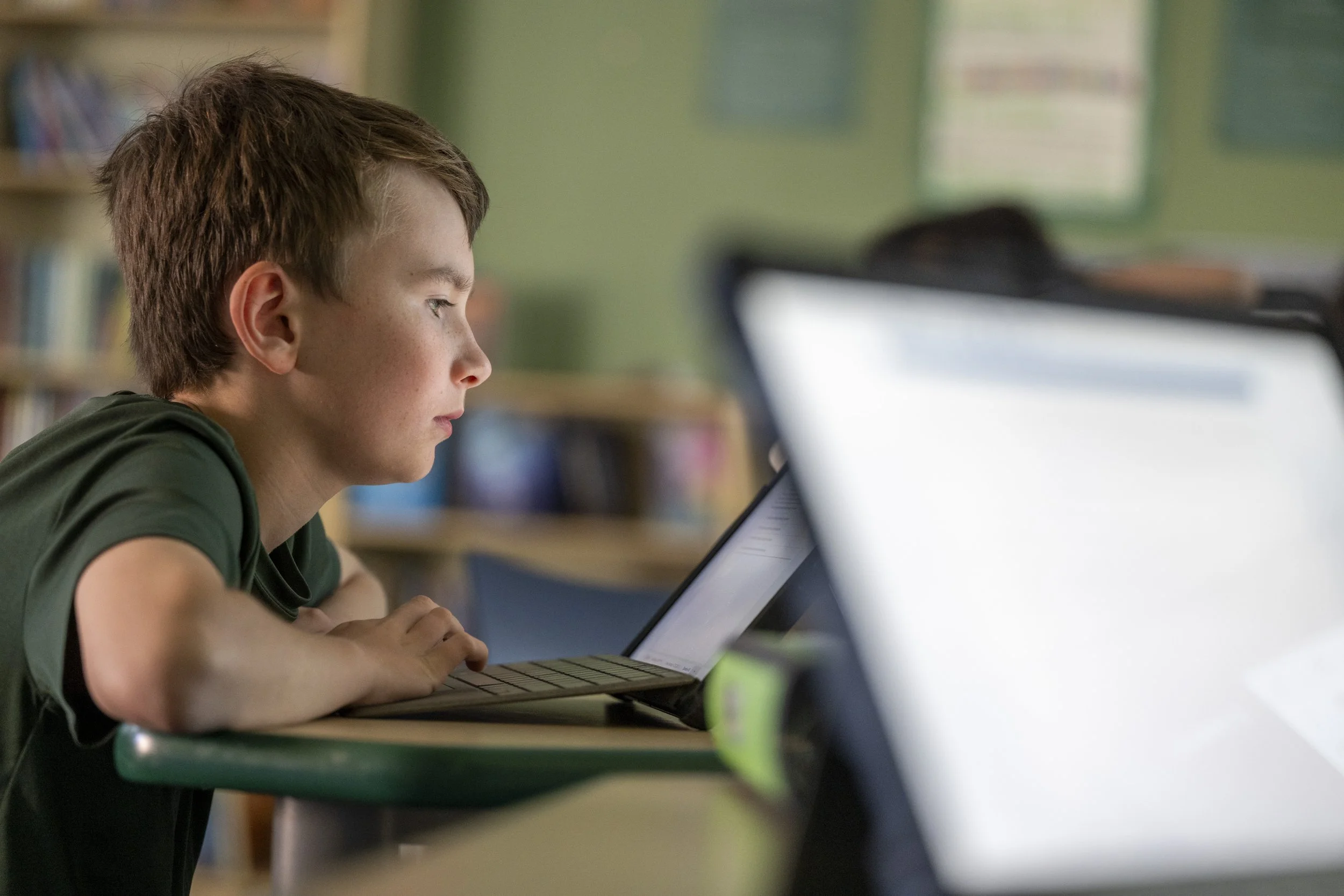 A young boy with brown hair studying at a school desk using a laptop, with bookshelves in the background.