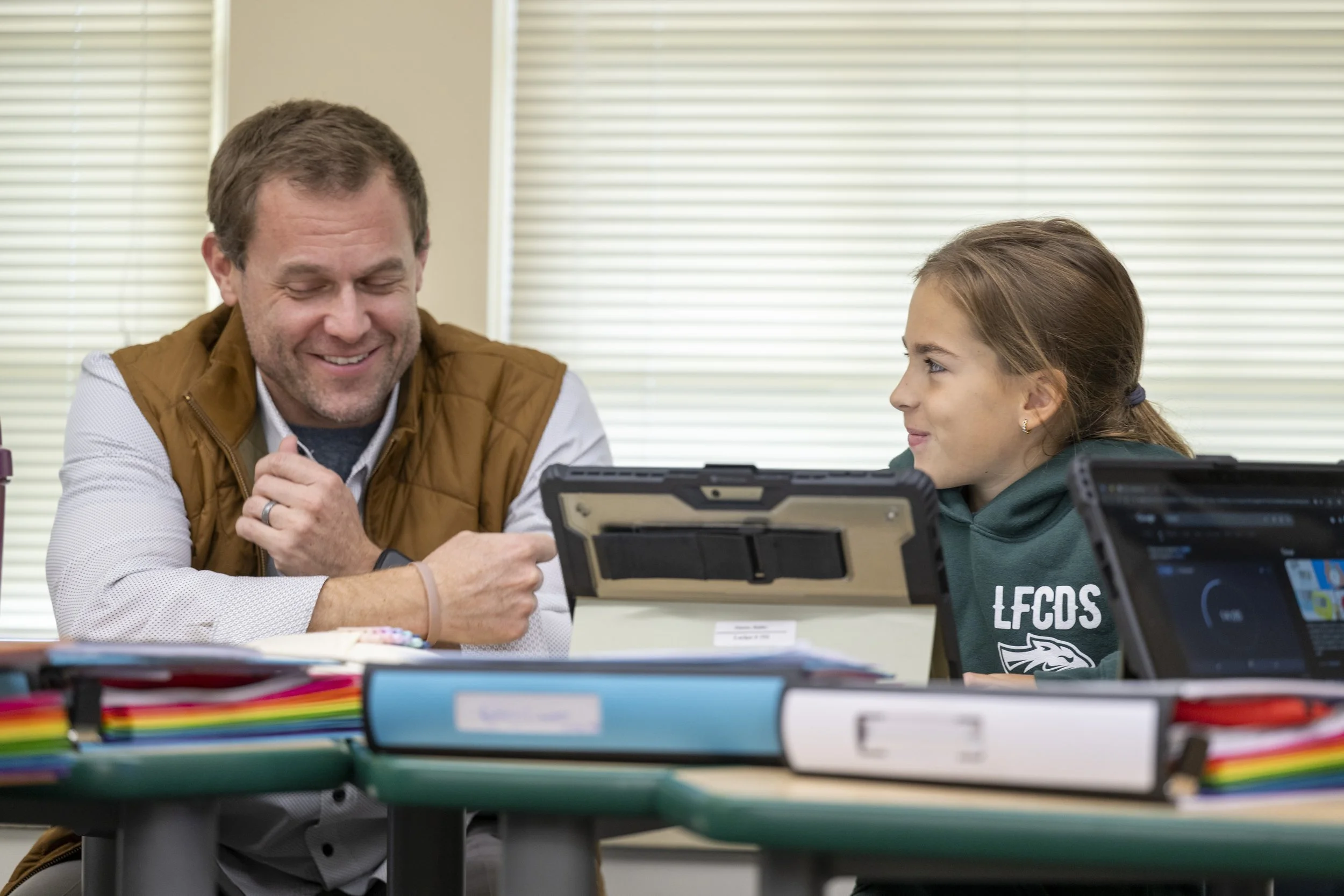 A man and a young girl sitting at a desk in a classroom, smiling and engaging in a learning activity with tablets and colorful school supplies around them.