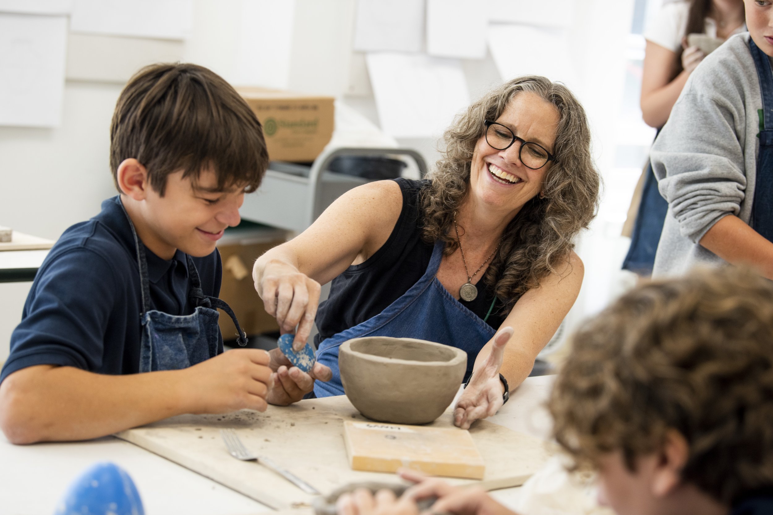 A woman and a young boy are working together on a pottery project in a classroom, smiling and focused on their clay pottery piece.