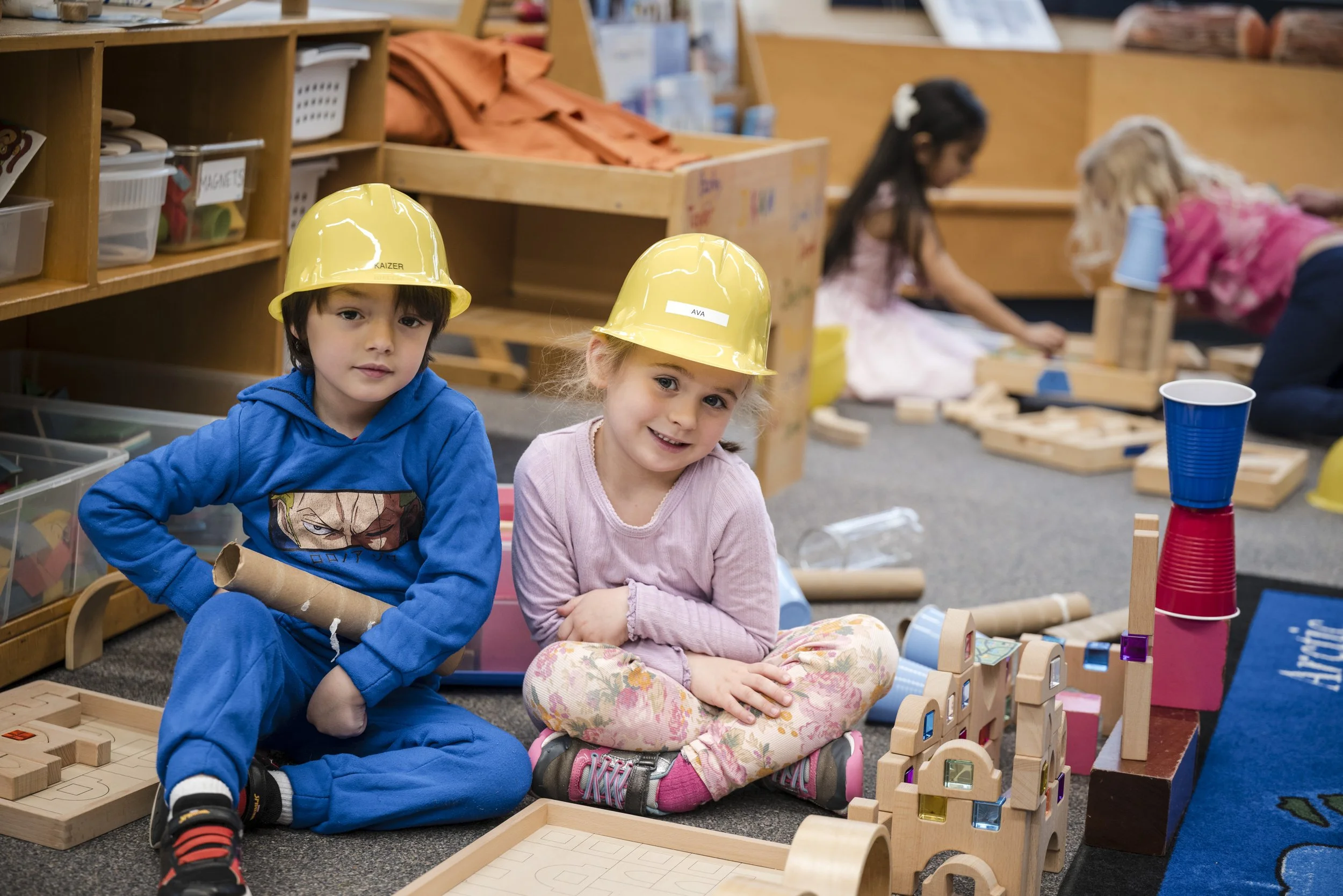 Children wearing yellow safety helmets sitting on the floor in a classroom, engaged in building with wooden blocks and toys.