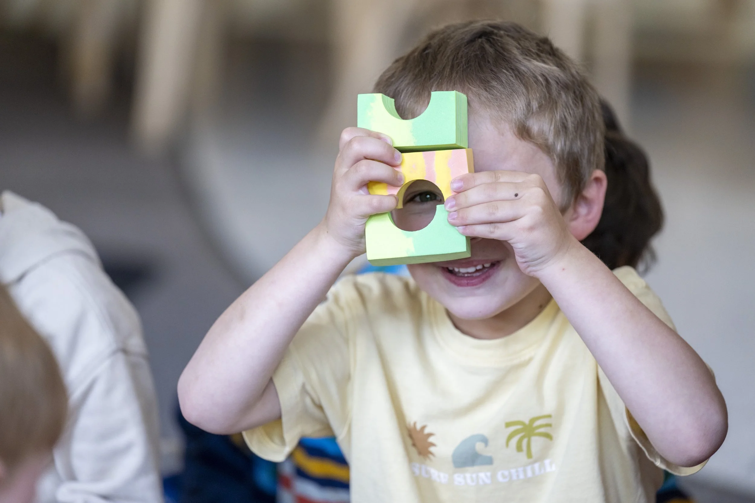 A young boy with curly hair holds a colorful sticky note pyramid up to his face, peeking through a hole in the middle, smiling and playing.